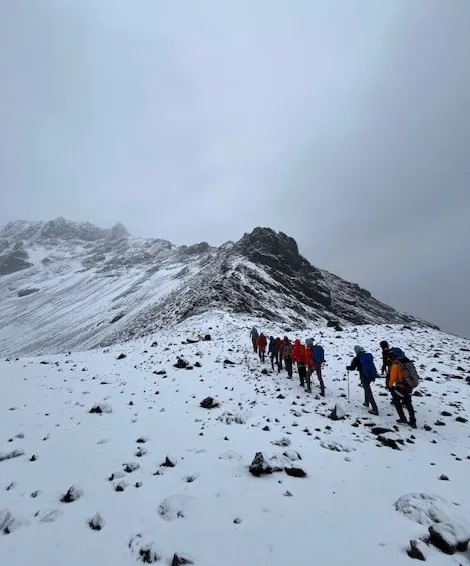 Group of hikers trekking up a snow-covered mountain slope under a cloudy sky.