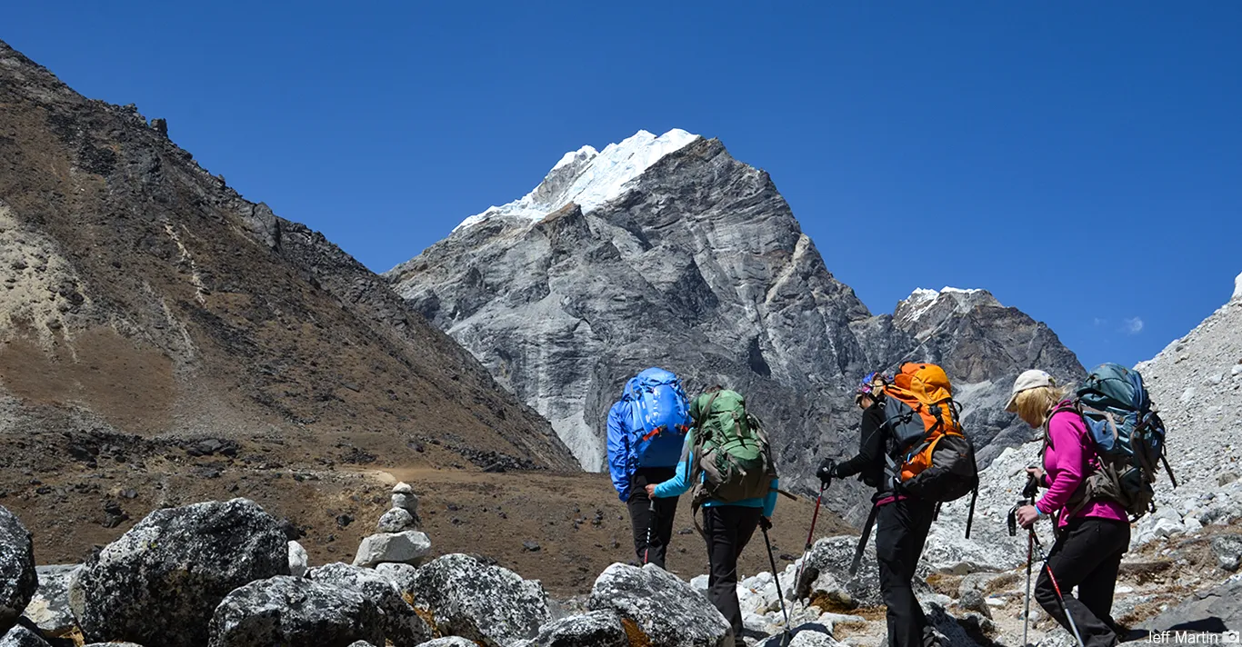 Four hikers with backpacks trekking rocky terrain toward a snow-capped mountain under clear blue sky.