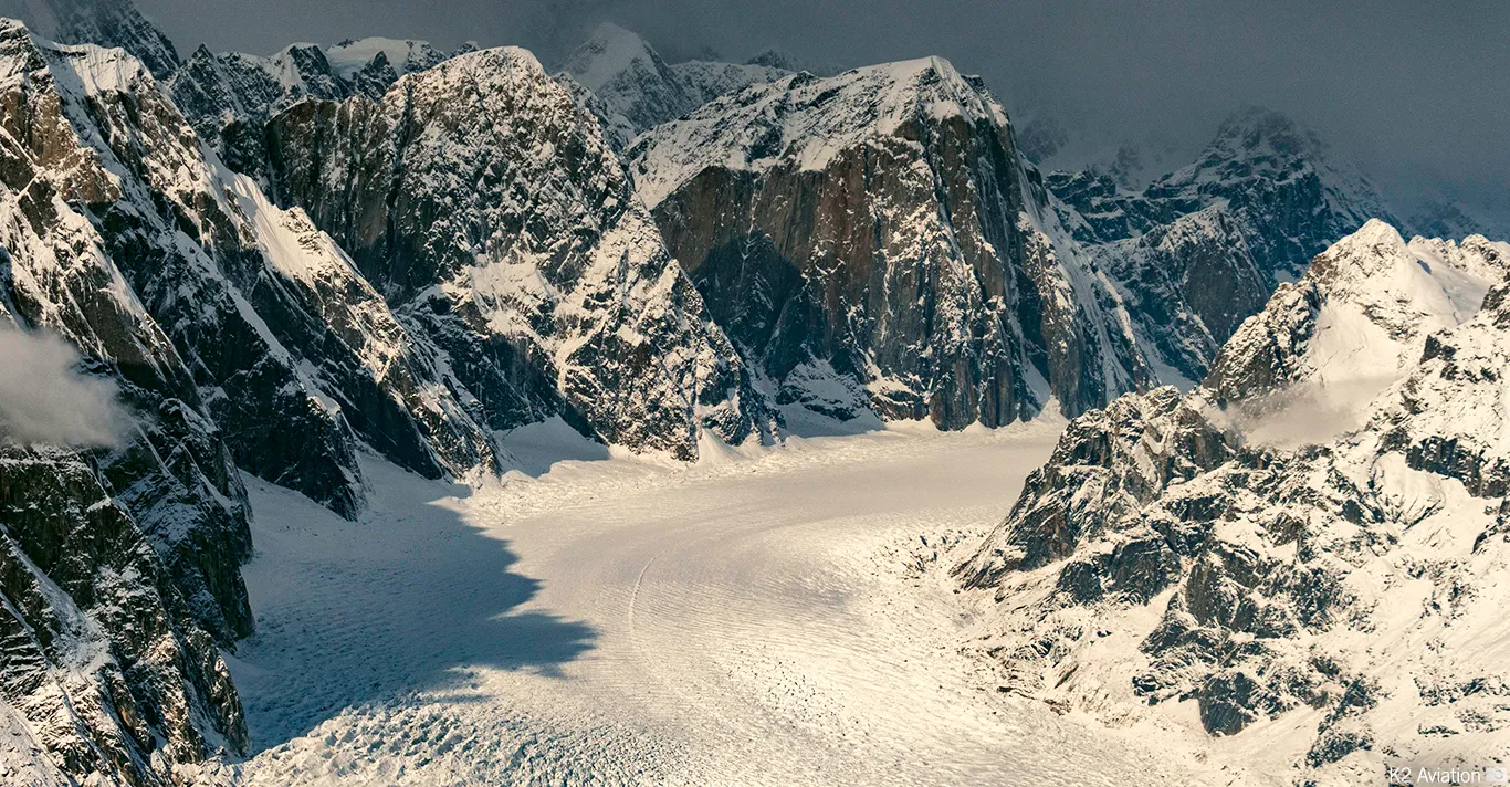 Snow-covered rugged mountain peaks surrounding a wide glacier under a cloudy sky.