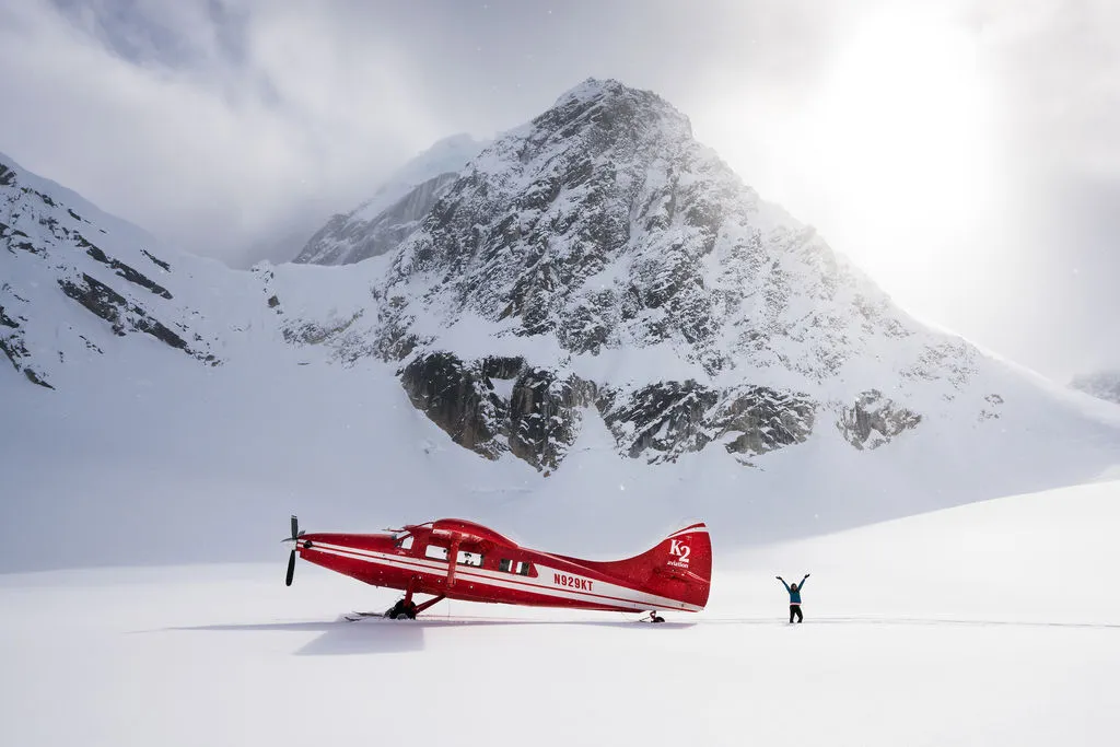 Red plane parked on snow with person raising arms in front of a large snowy mountain.