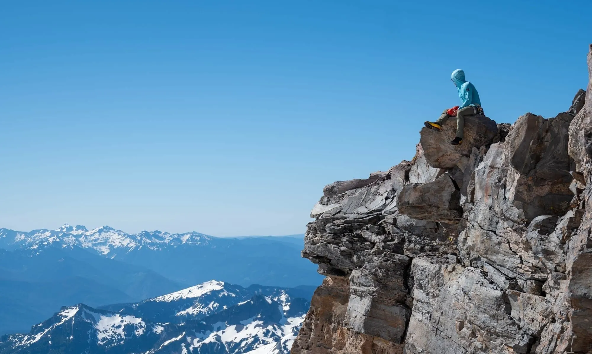 Person in blue hoodie sitting on a rocky cliff with snowy mountains and clear blue sky in the background.