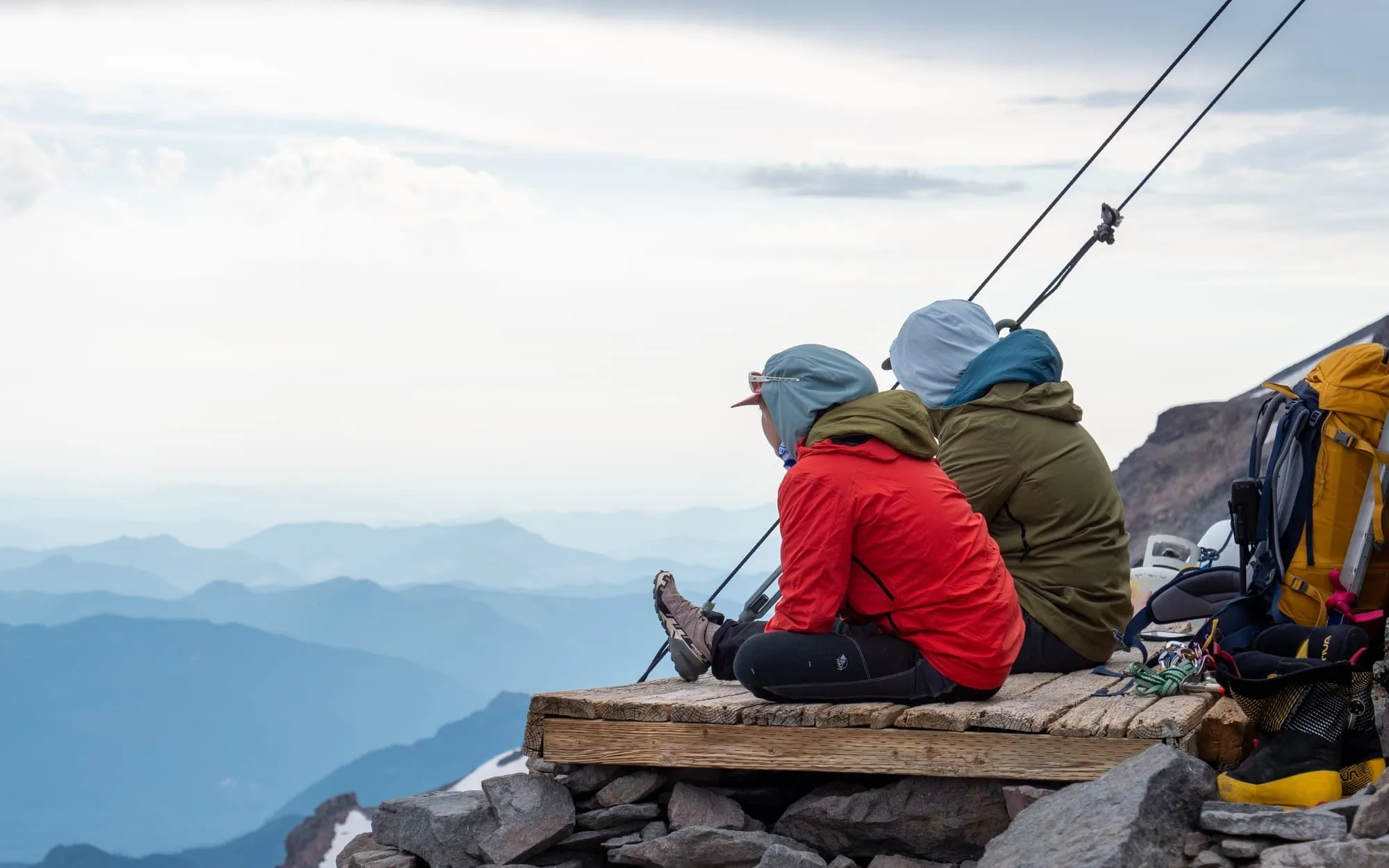 Two hikers sitting on a wooden platform overlooking misty mountain layers under a cloudy sky.