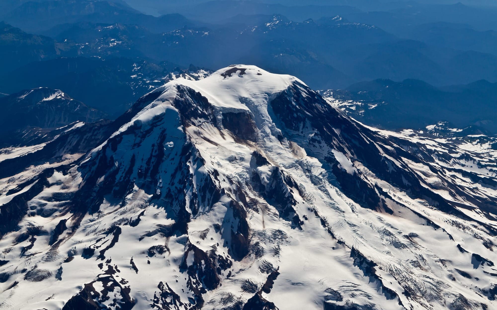Aerial view of snow-covered mountain peak with surrounding rugged terrain and blue distant mountains.