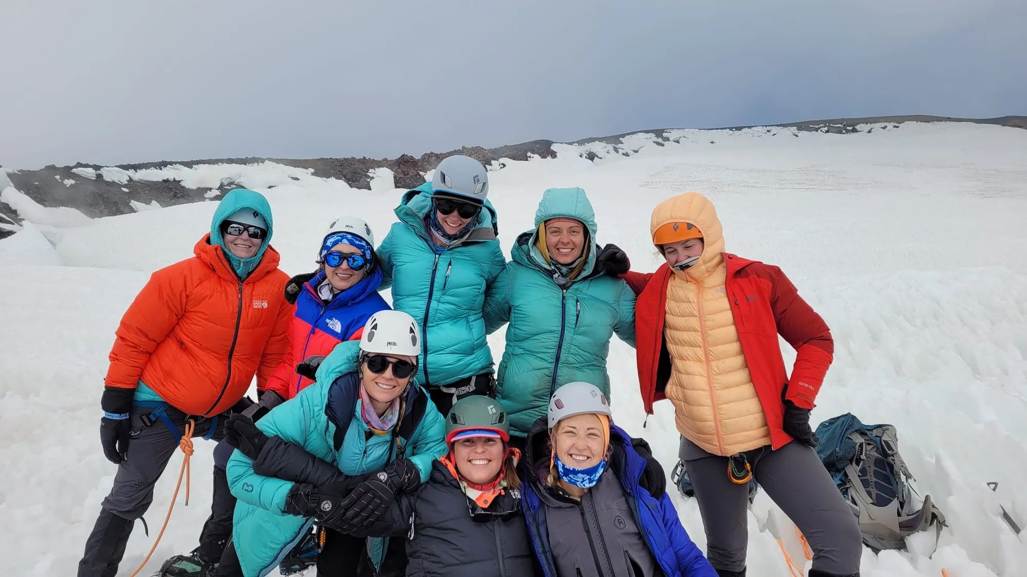 Group of eight women climbers in winter gear posing happy on a snowy mountain glacier.