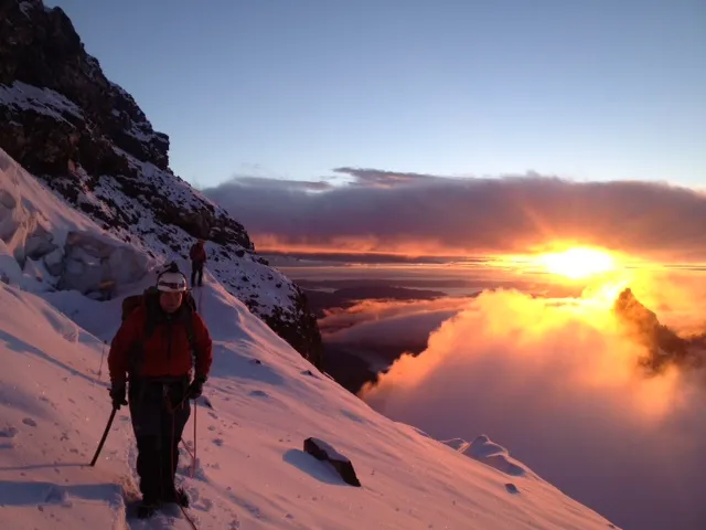 Two climbers descend a snowy mountain slope during a sunrise with clouds and peaks in the background.