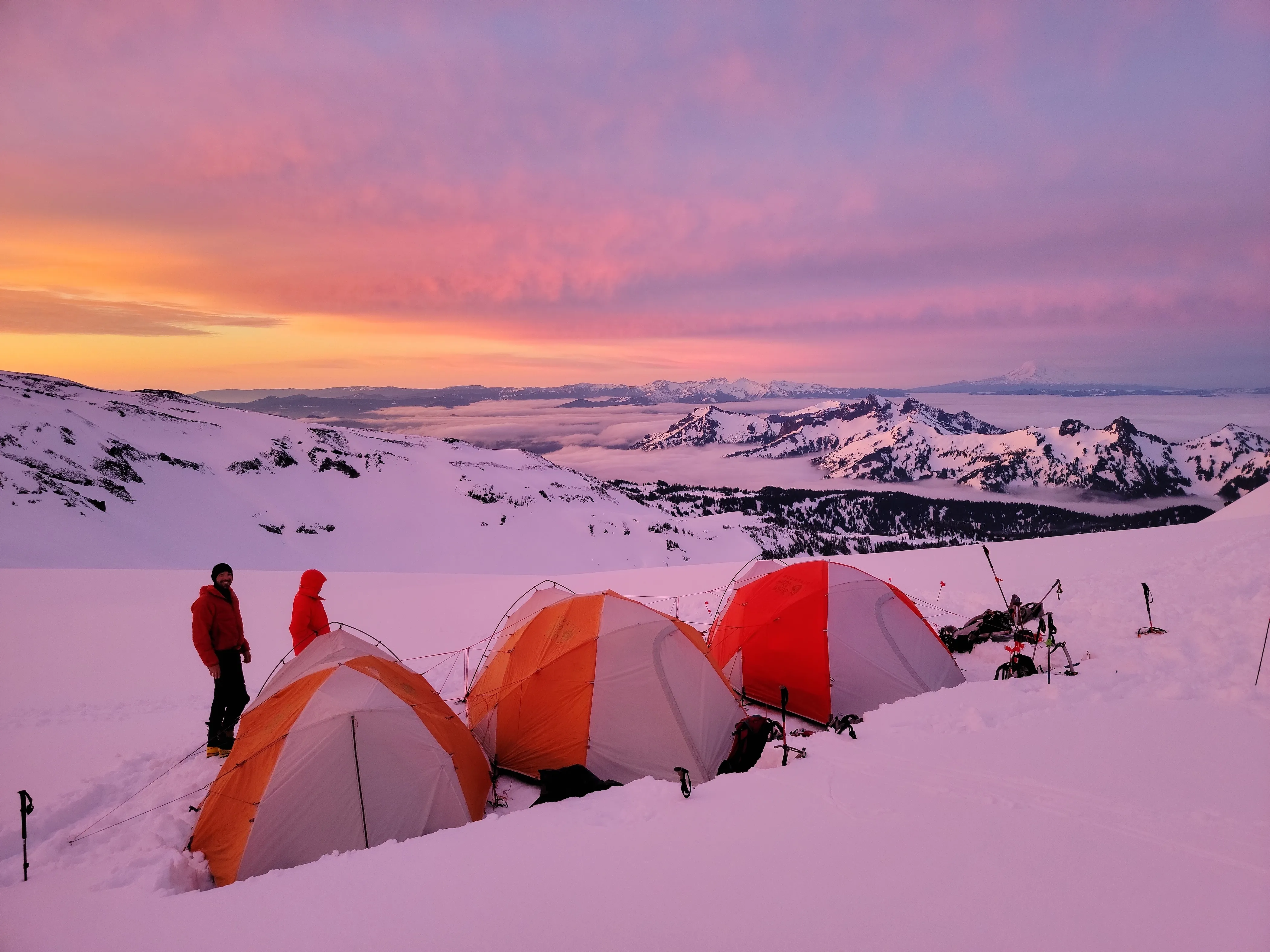 Three orange tents on snow with two people nearby at sunset in a snowy mountain landscape.