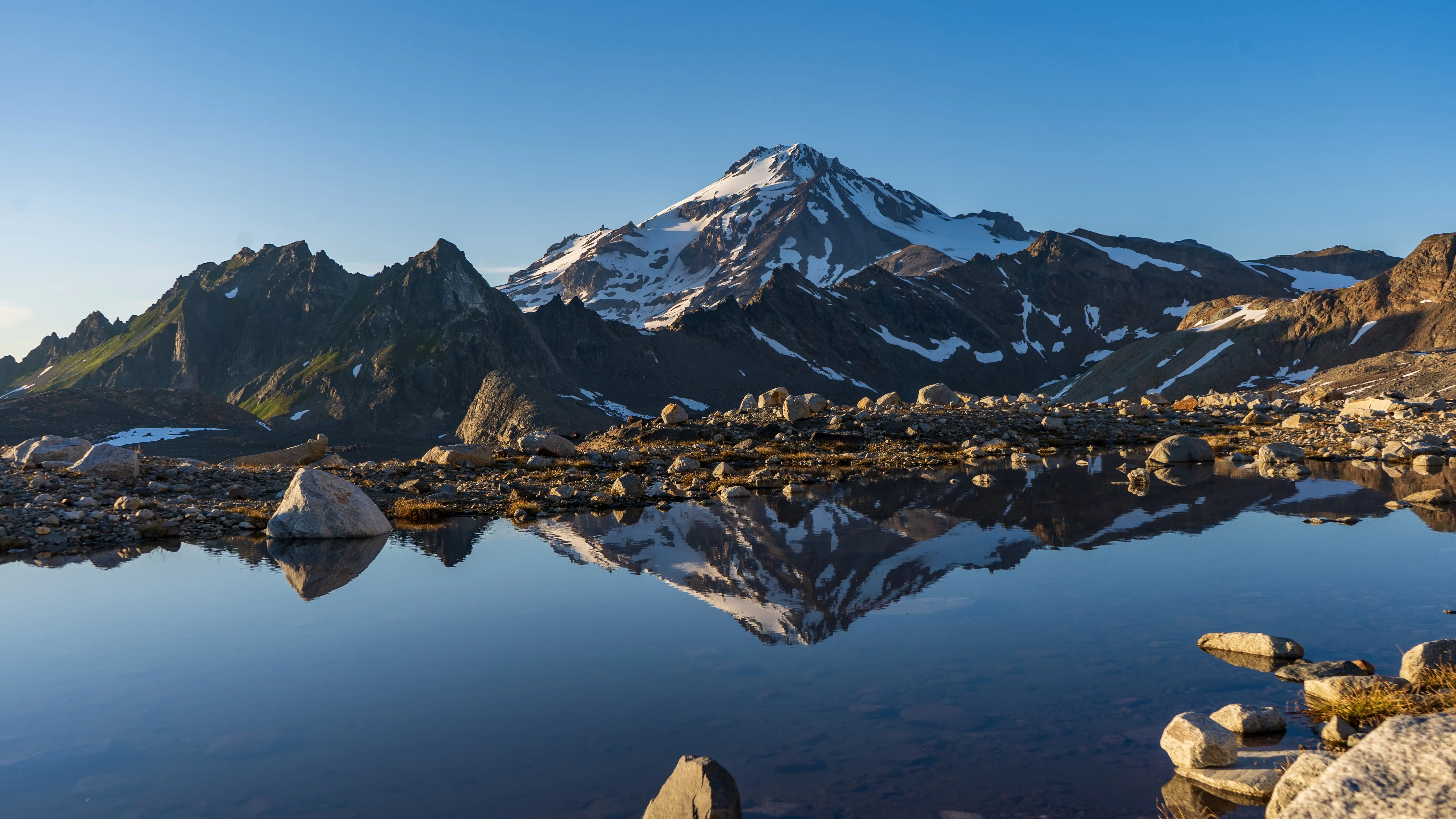 Snow-capped mountain and peaks reflected in a calm alpine lake surrounded by rocks.