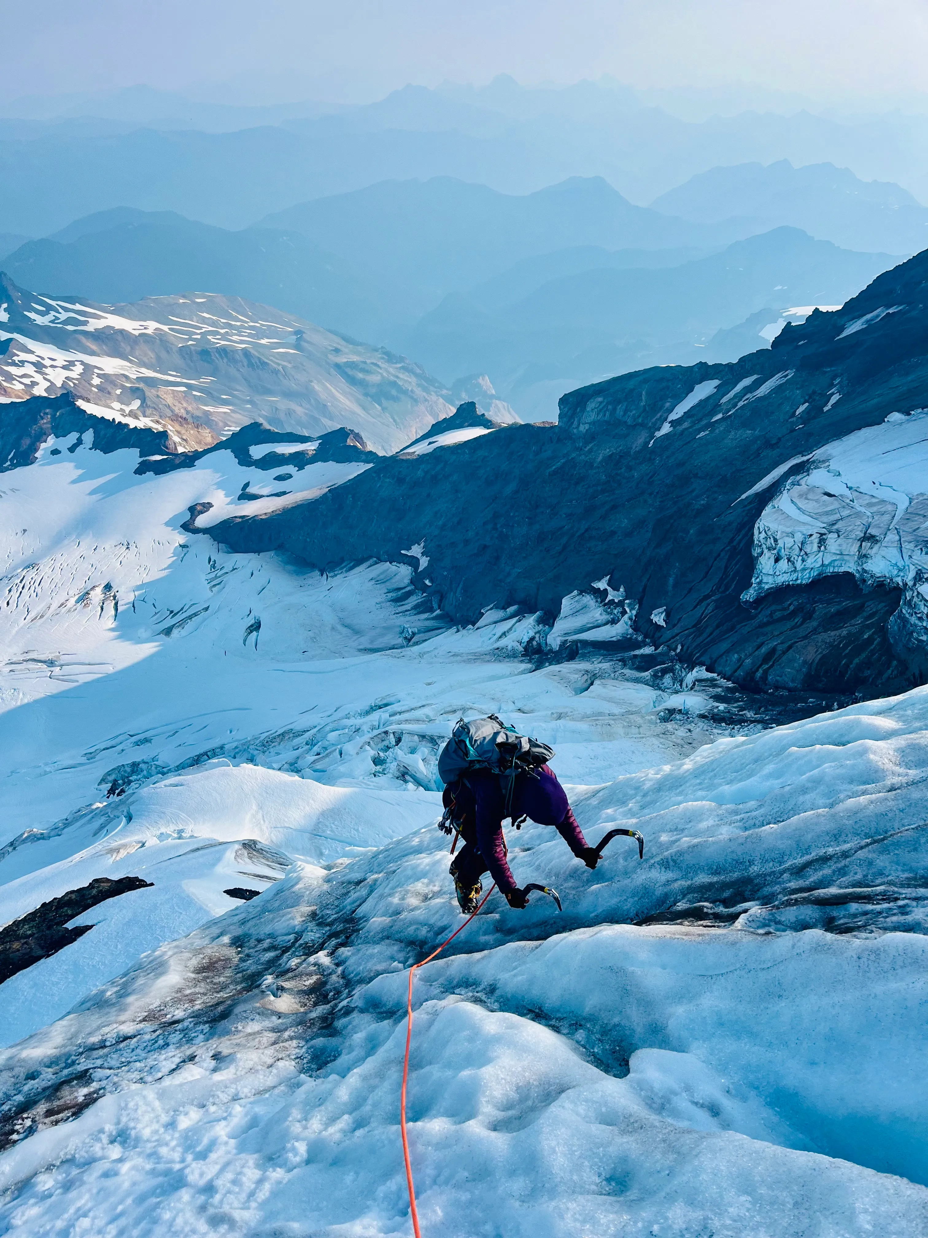 Climber with ice axes ascending a snowy glacier mountain with a rope trailing behind.