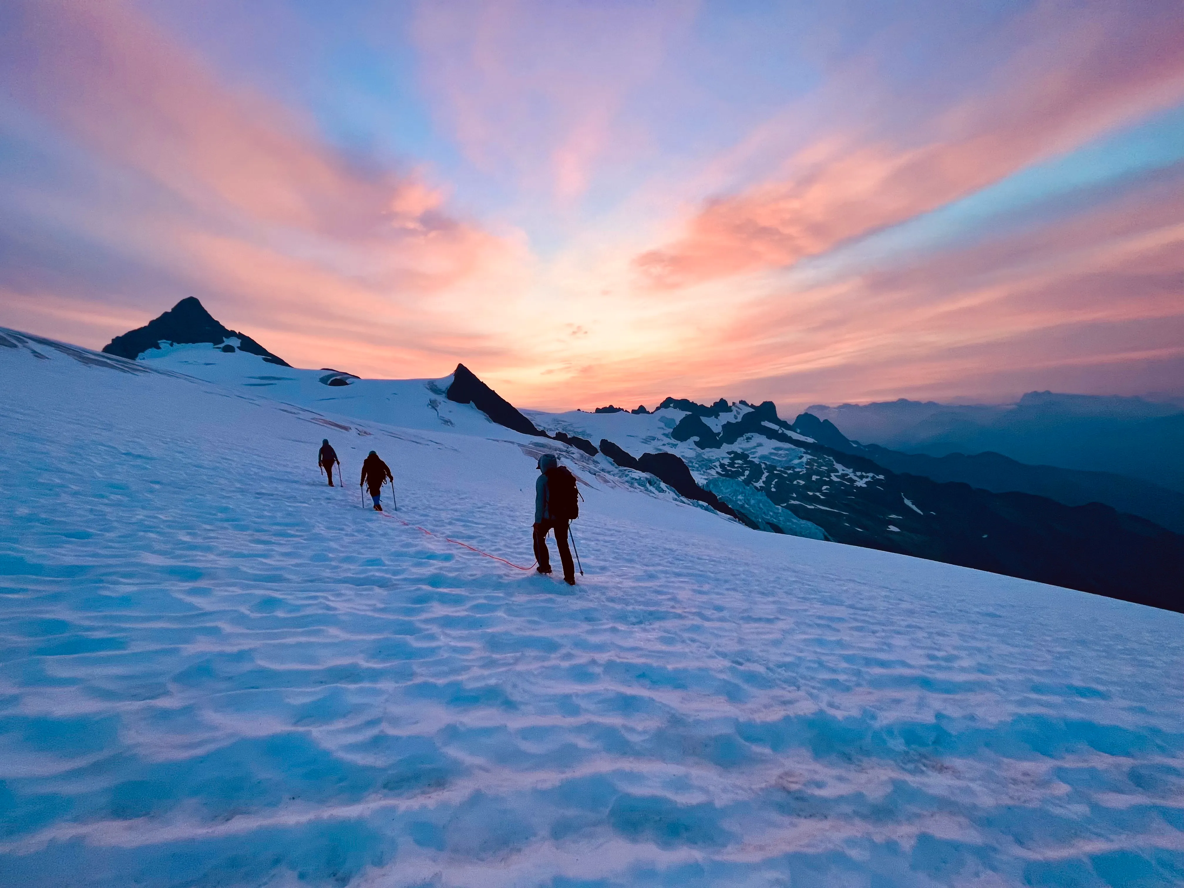 Three climbers roped together ascending snowy mountain slope at vibrant sunrise or sunset.