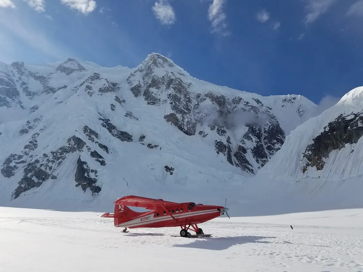 Red K2 airplane parked on snowfield in front of tall snowy mountain under blue sky.