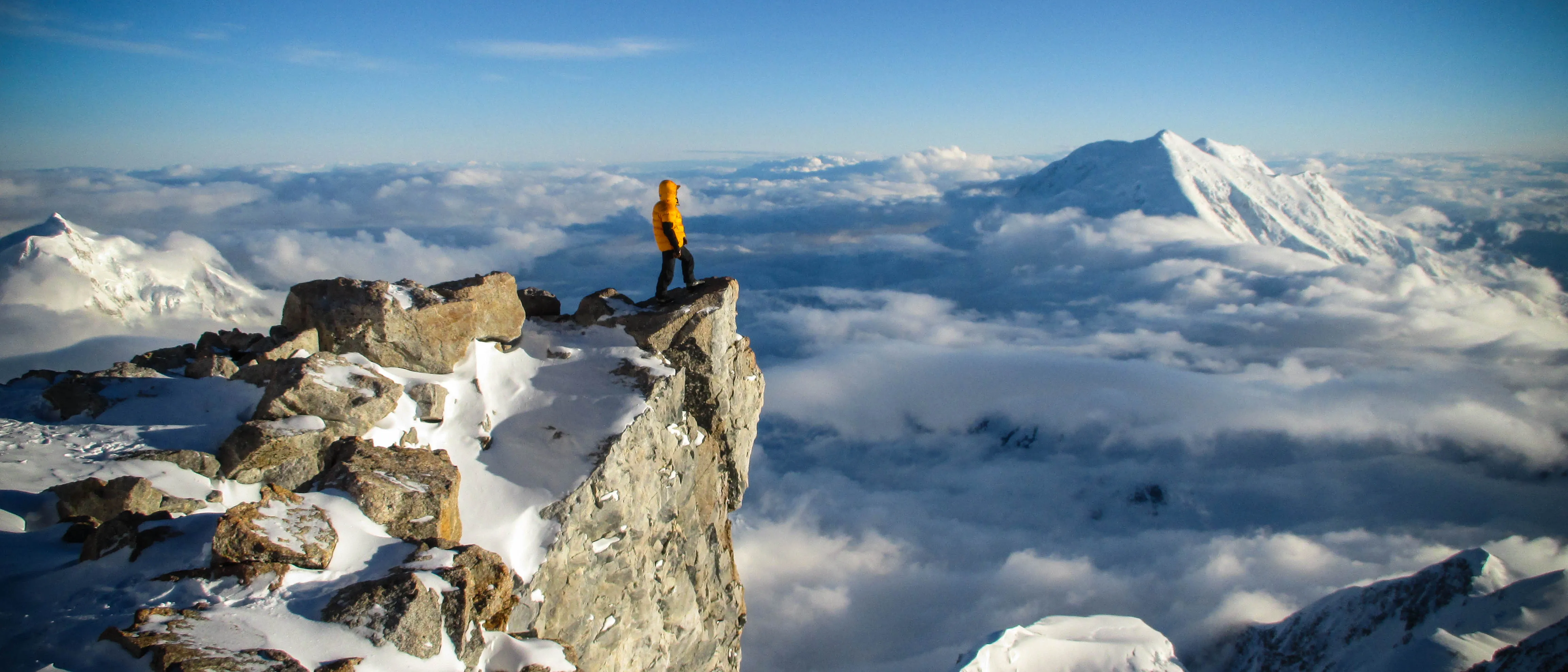 Person in a yellow jacket stands on snowy mountain cliff overlooking clouds and distant peaks.