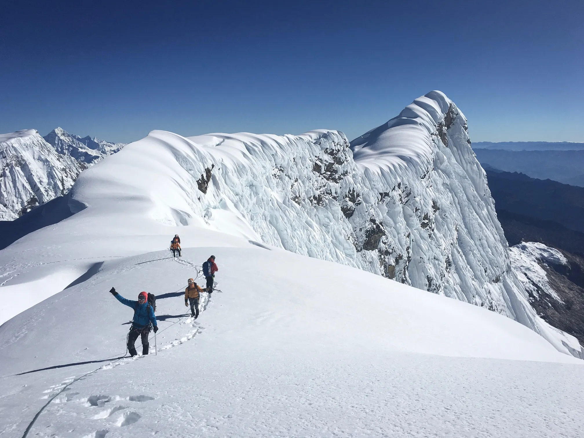 Four mountaineers climbing a snowy ridge with clear skies and rugged mountain peaks in the background.