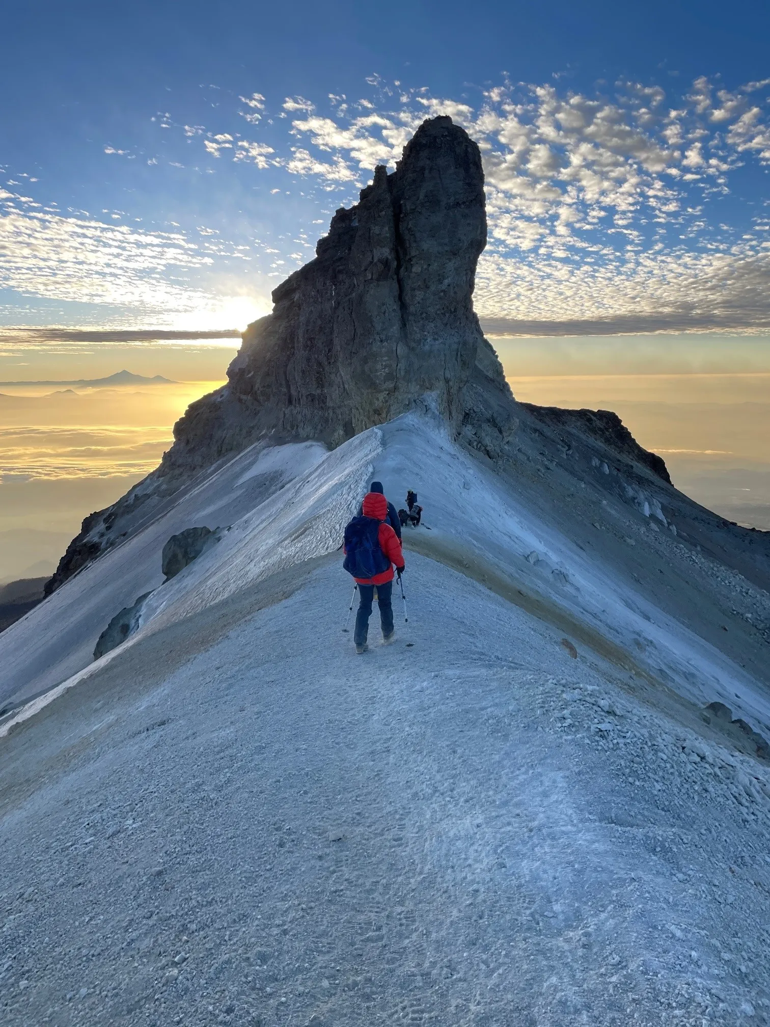 Hikers climbing a snowy ridge toward a tall rocky peak at sunrise with a cloudy sky.