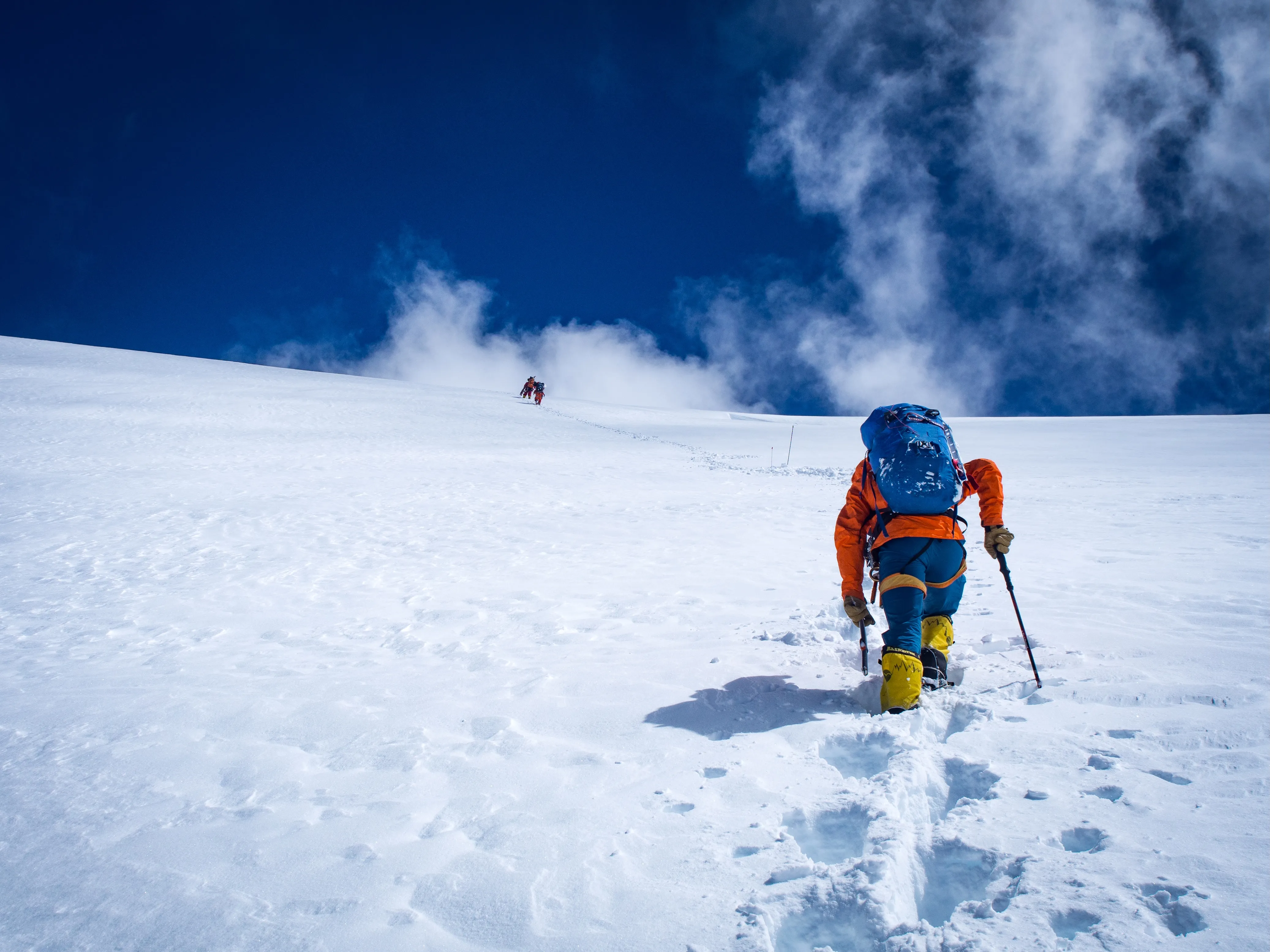 Climbers in orange jackets hiking up a snowy mountain under a deep blue sky with clouds.