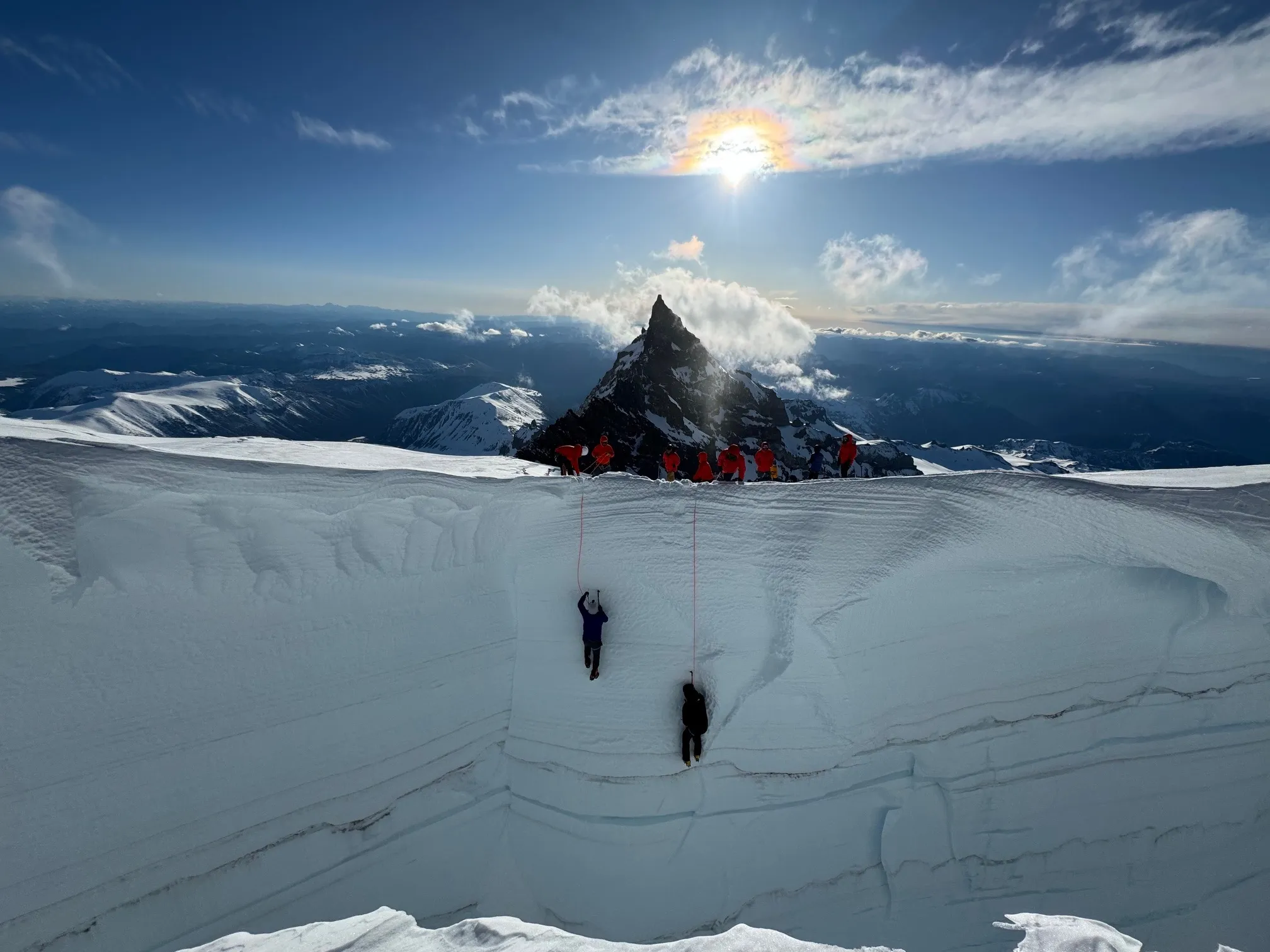 Climbers in red jackets assist two people climbing a steep snowy crevasse on a mountain peak.
