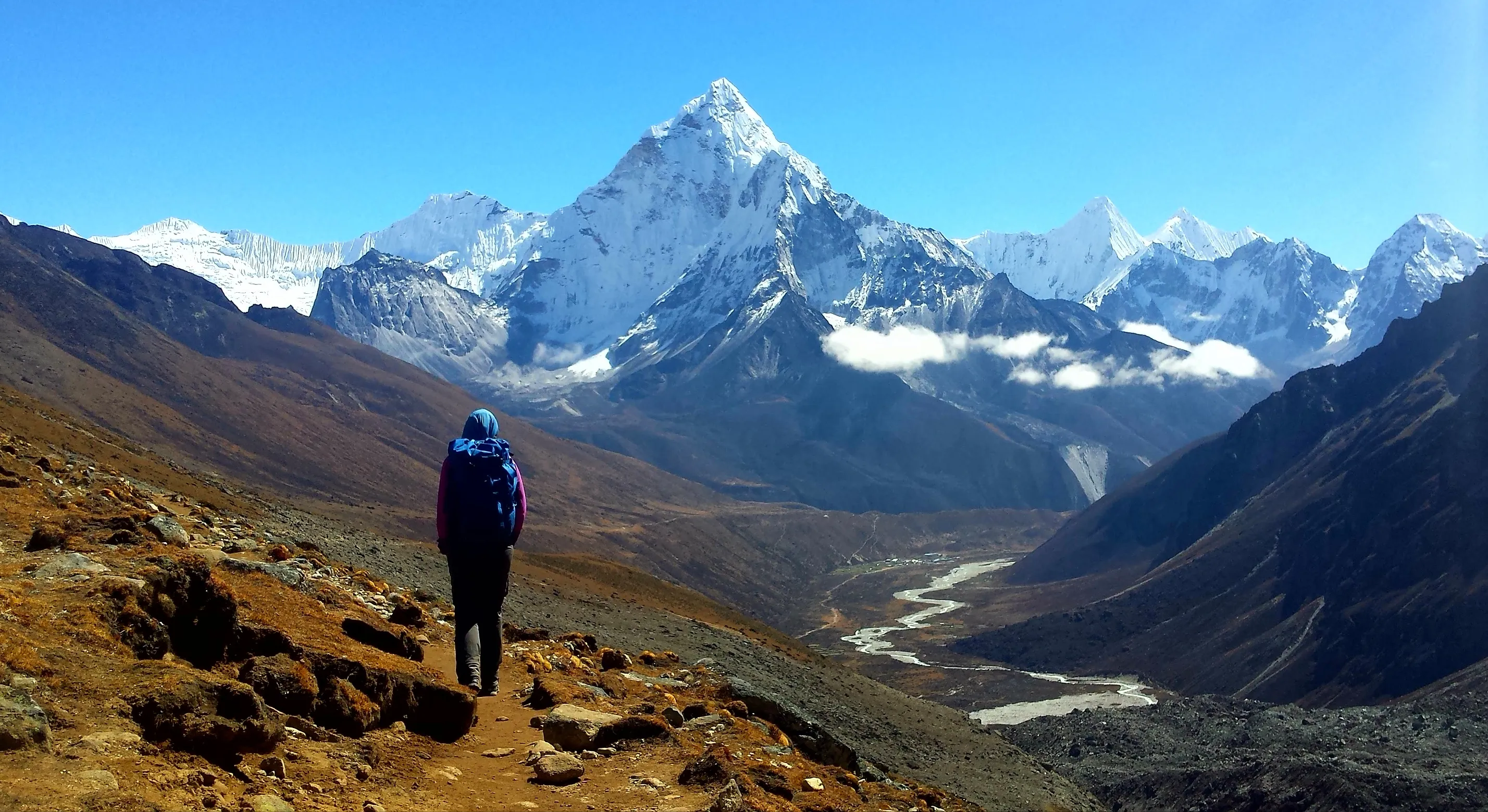 Person hiking on mountain trail with snow-covered peaks and winding river in a clear blue sky.