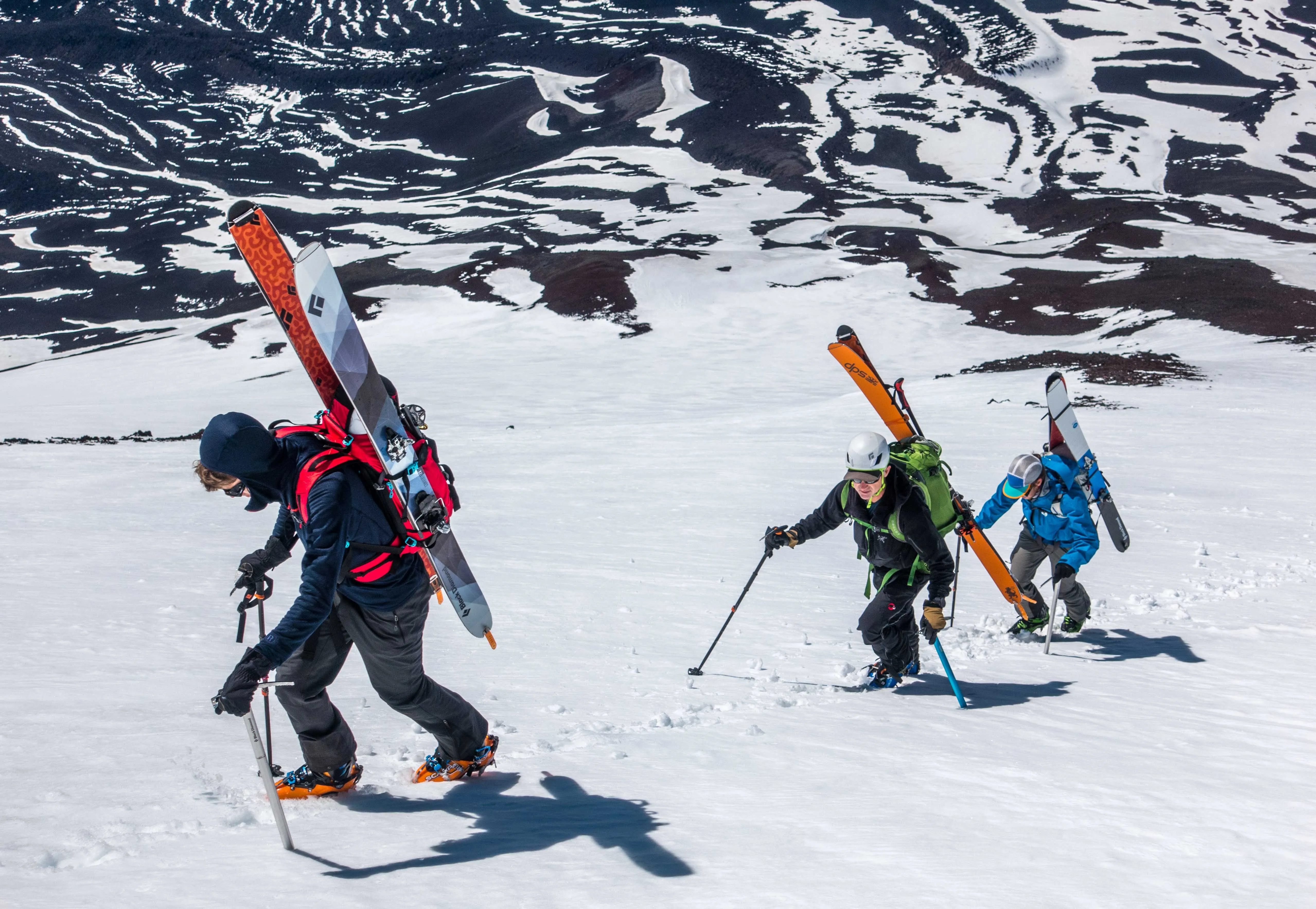 Three climbers hiking up a snowy mountain carrying skis and wearing backpacks and helmets.
