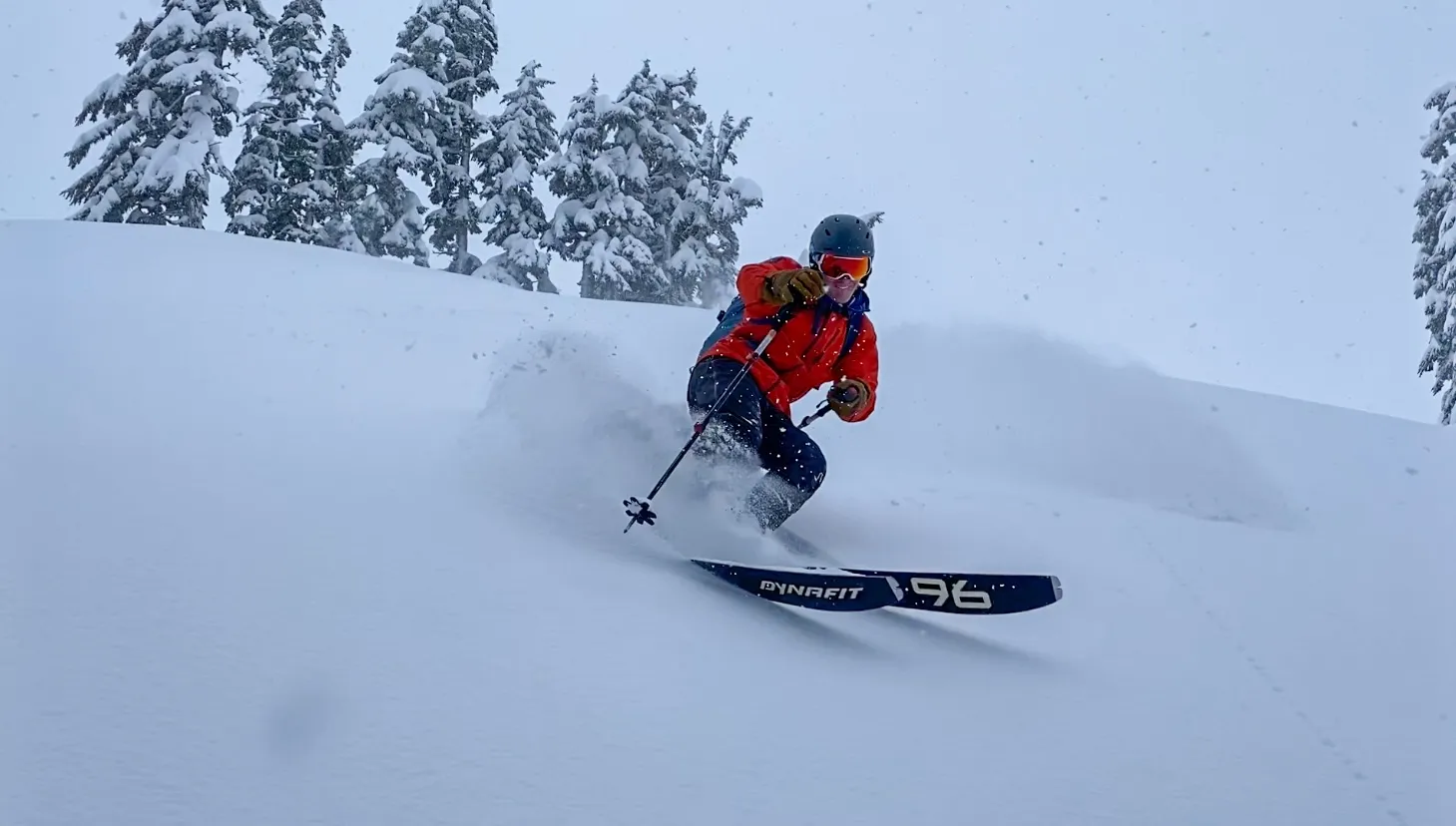 Skier in orange jacket and helmet making a turn in fresh powder snow with snowy trees in background.