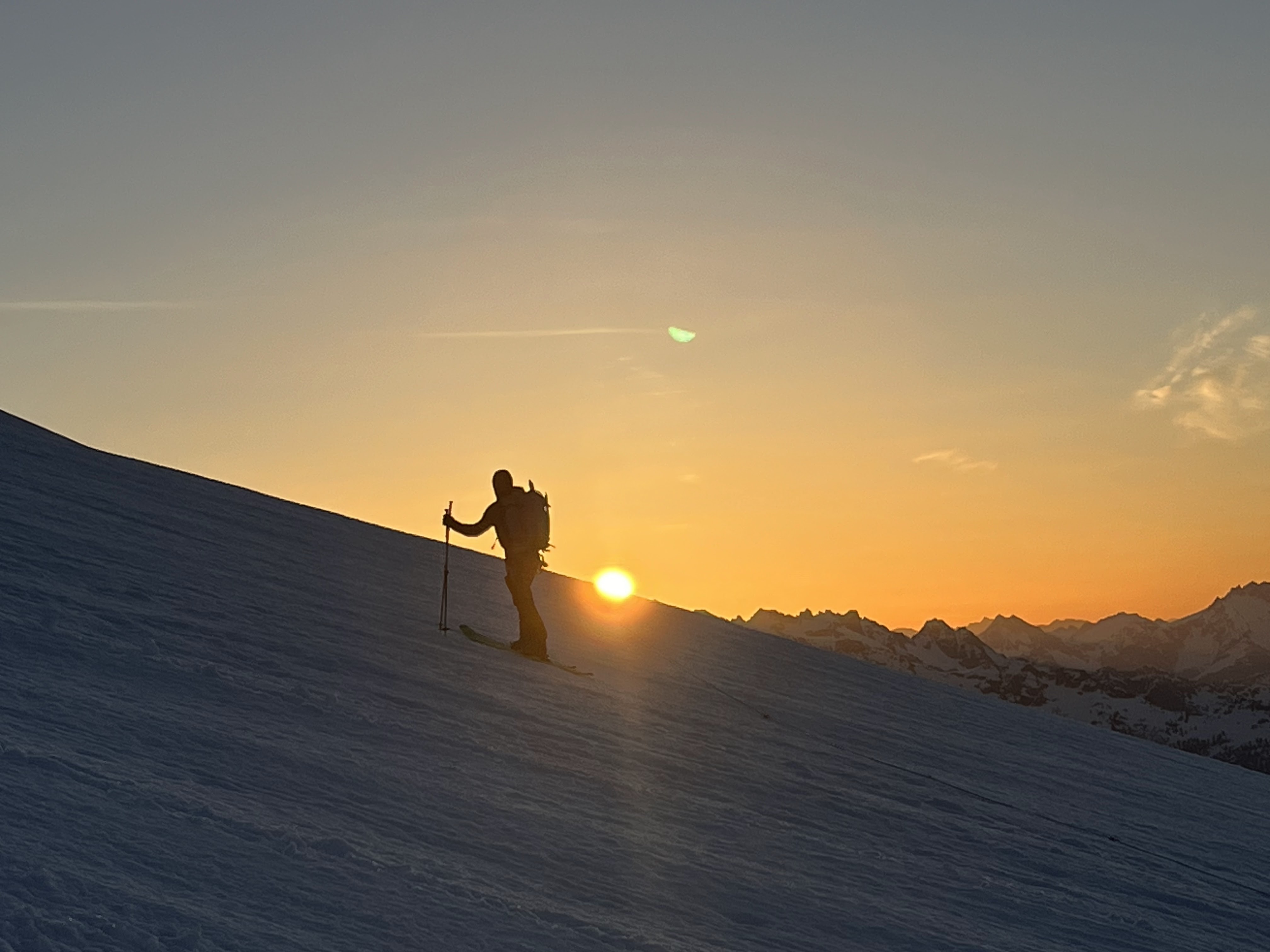Silhouette of skier ascending a snowy slope with hiking poles at sunrise over mountain peaks.