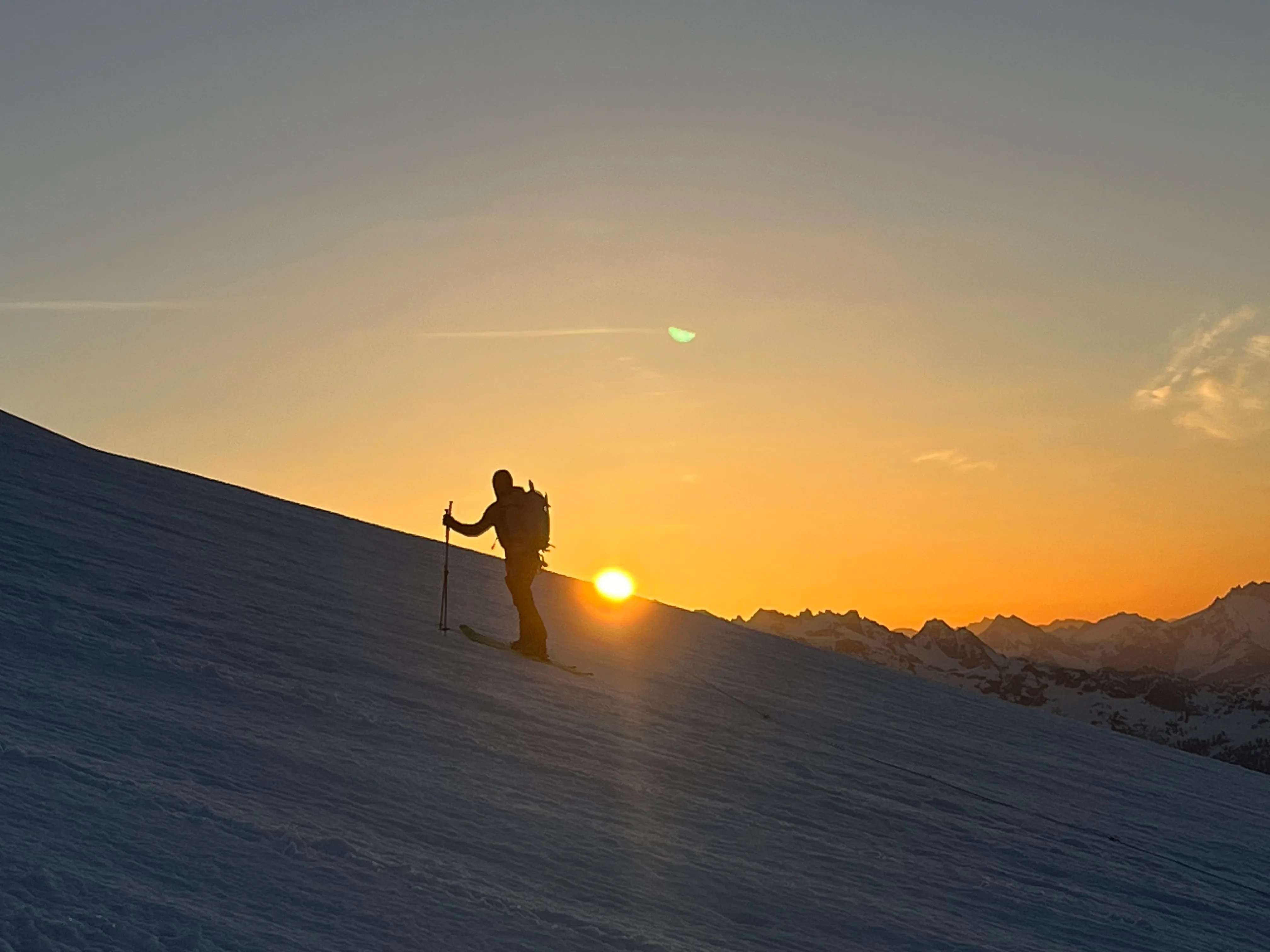 Silhouette of skier ascending a snowy slope with hiking poles at sunrise over mountain peaks.