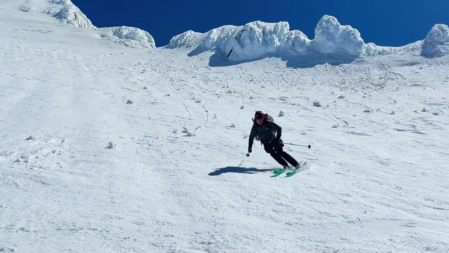 Skier in dark clothing with green skis carving turns down a snowy mountain slope under clear blue sky.