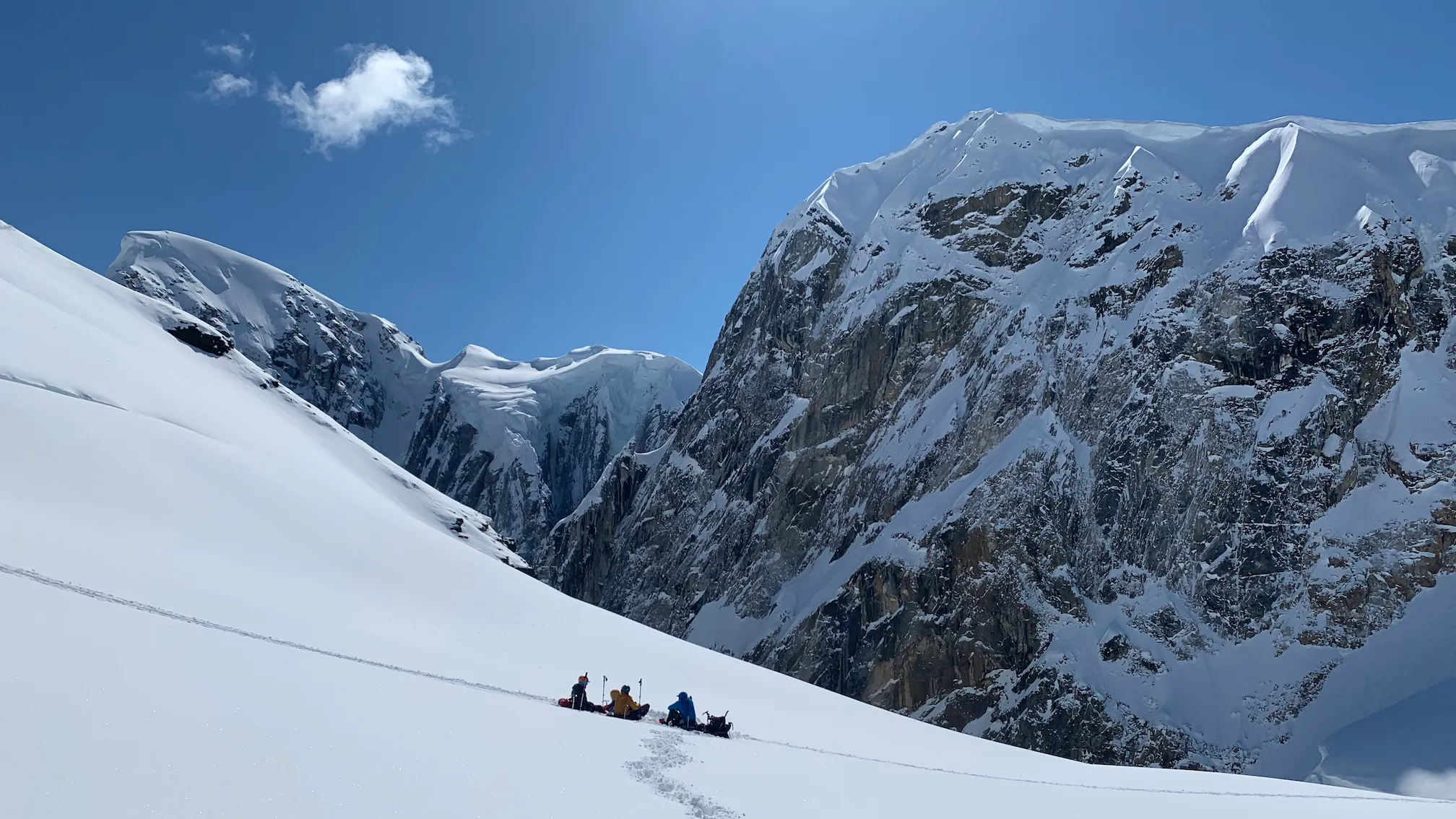 Four climbers resting on snowy mountain slope under clear blue sky with steep snow-covered cliffs.