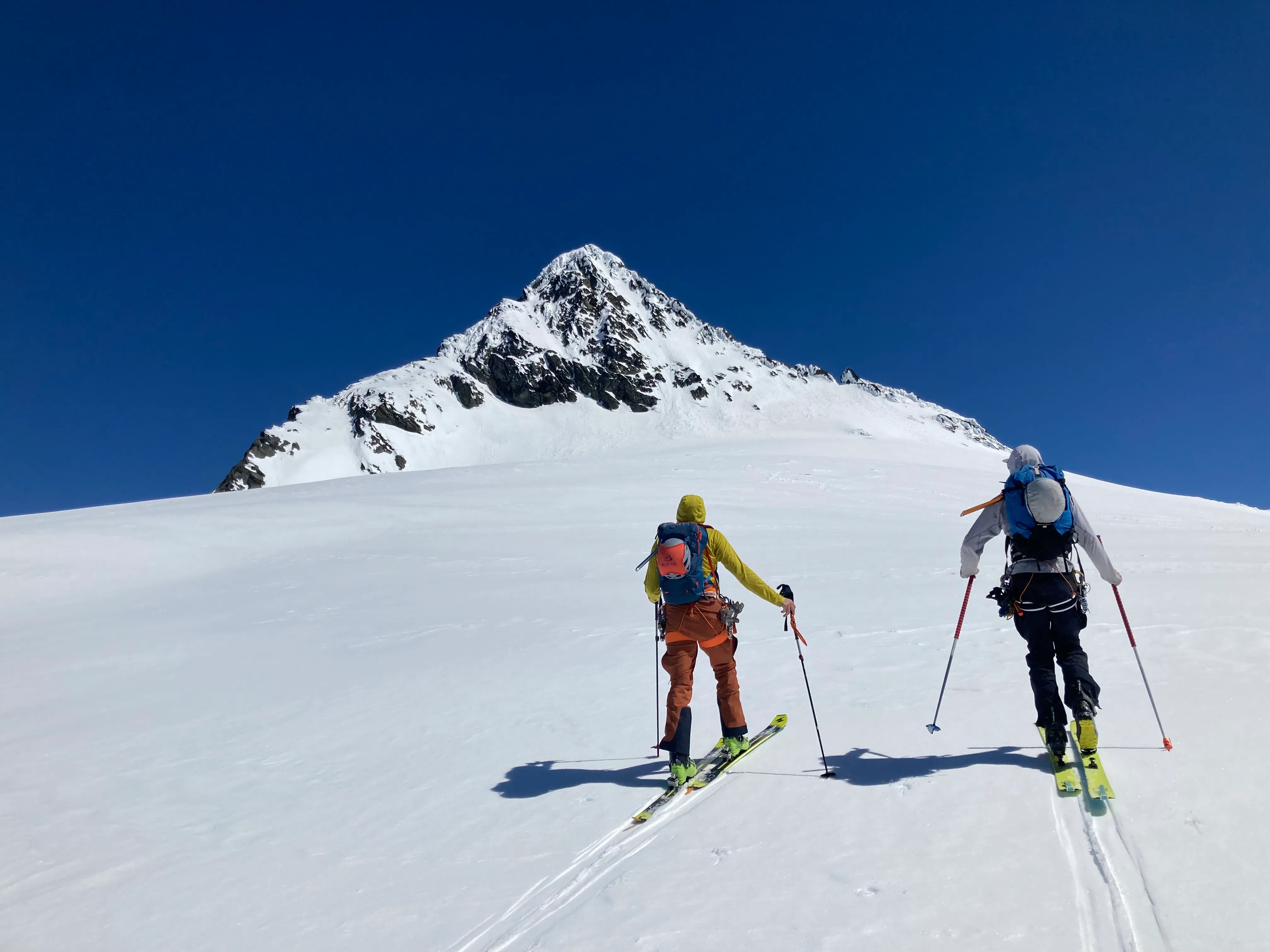 Two skiers ascending snowy mountain slope with a steep, rocky peak under clear blue sky.