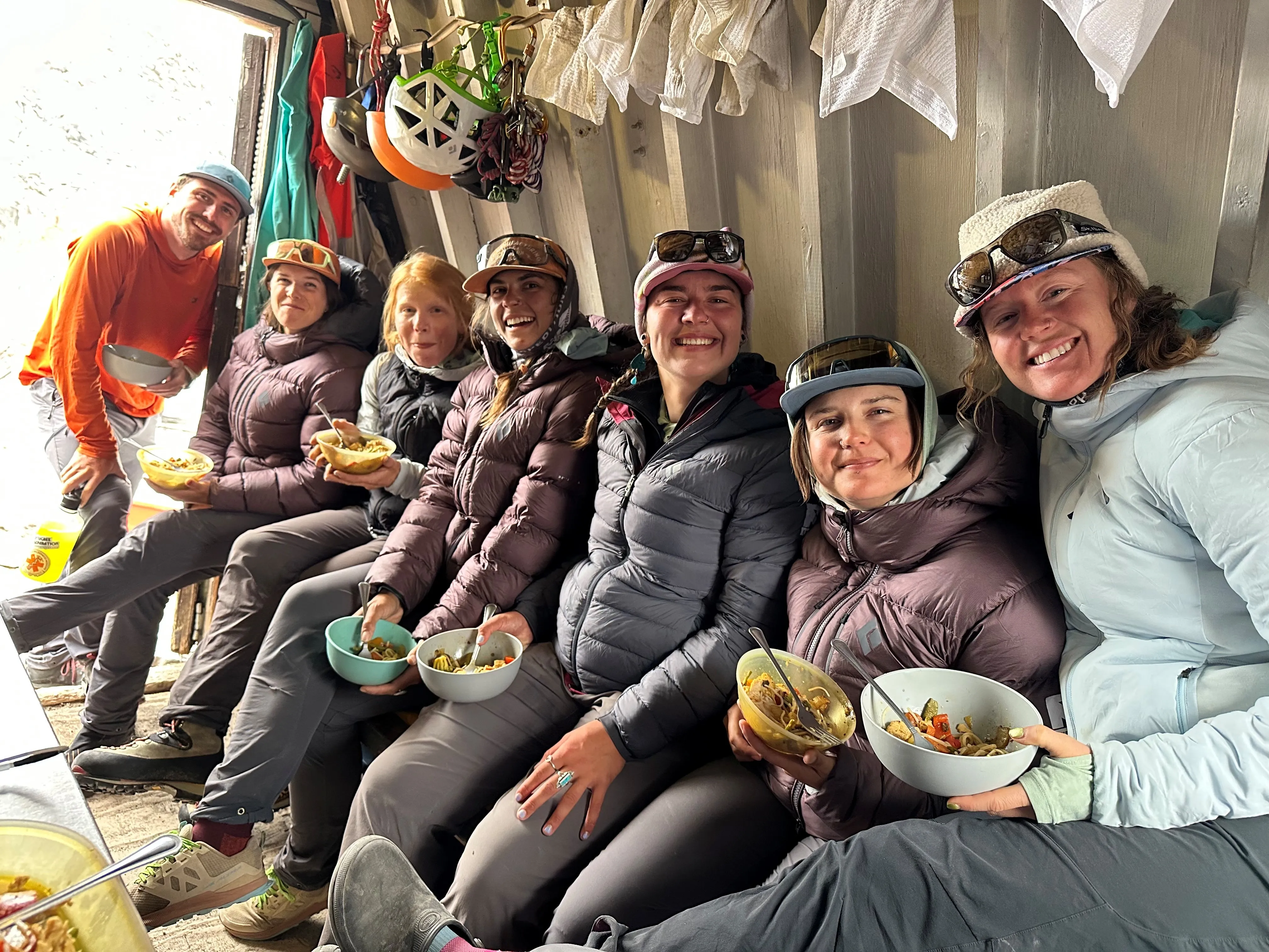 Group of seven people in outdoor gear sitting indoors eating from bowls and smiling at the camera.