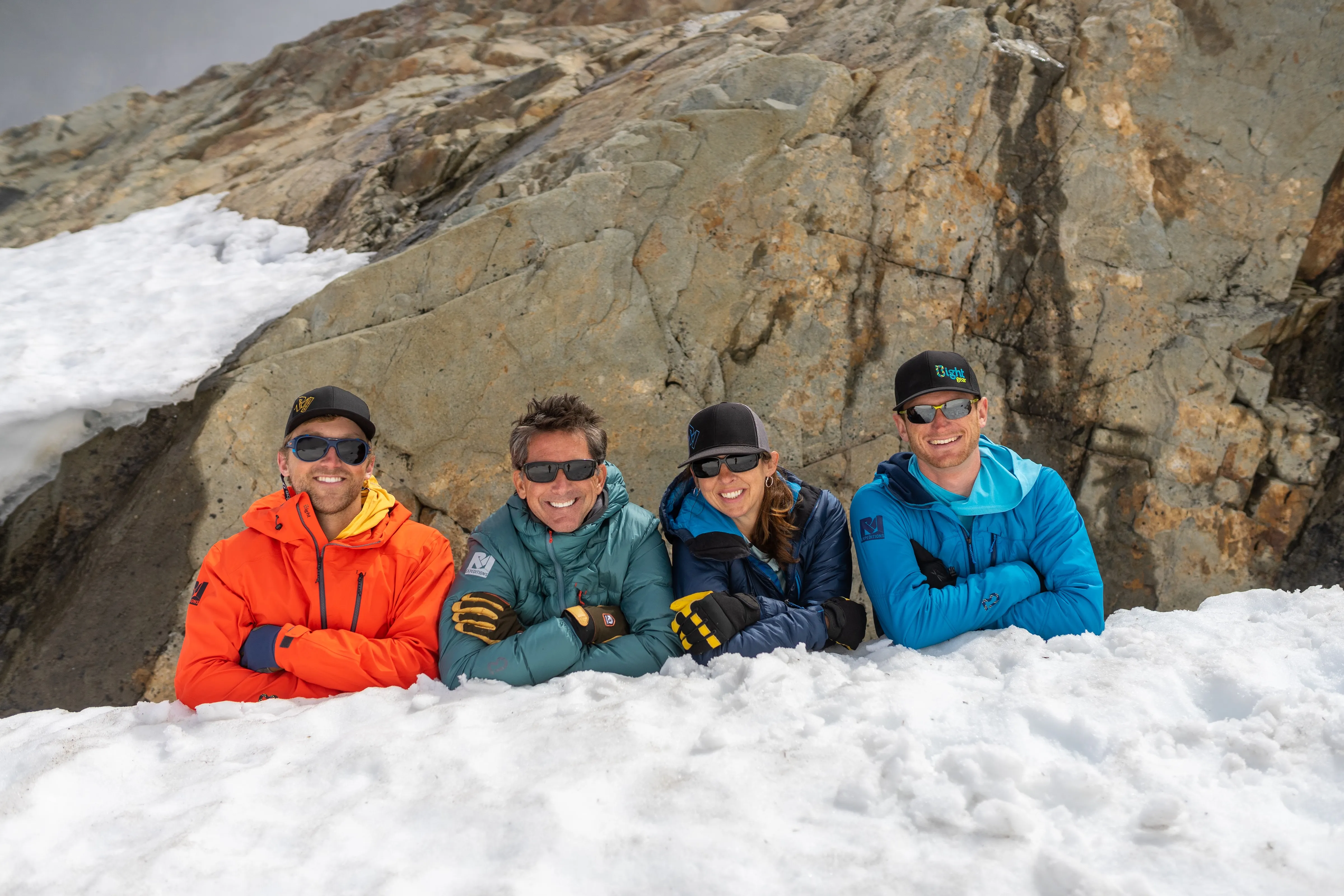Four people in colorful winter jackets and sunglasses smiling behind snow with rocky mountain background.