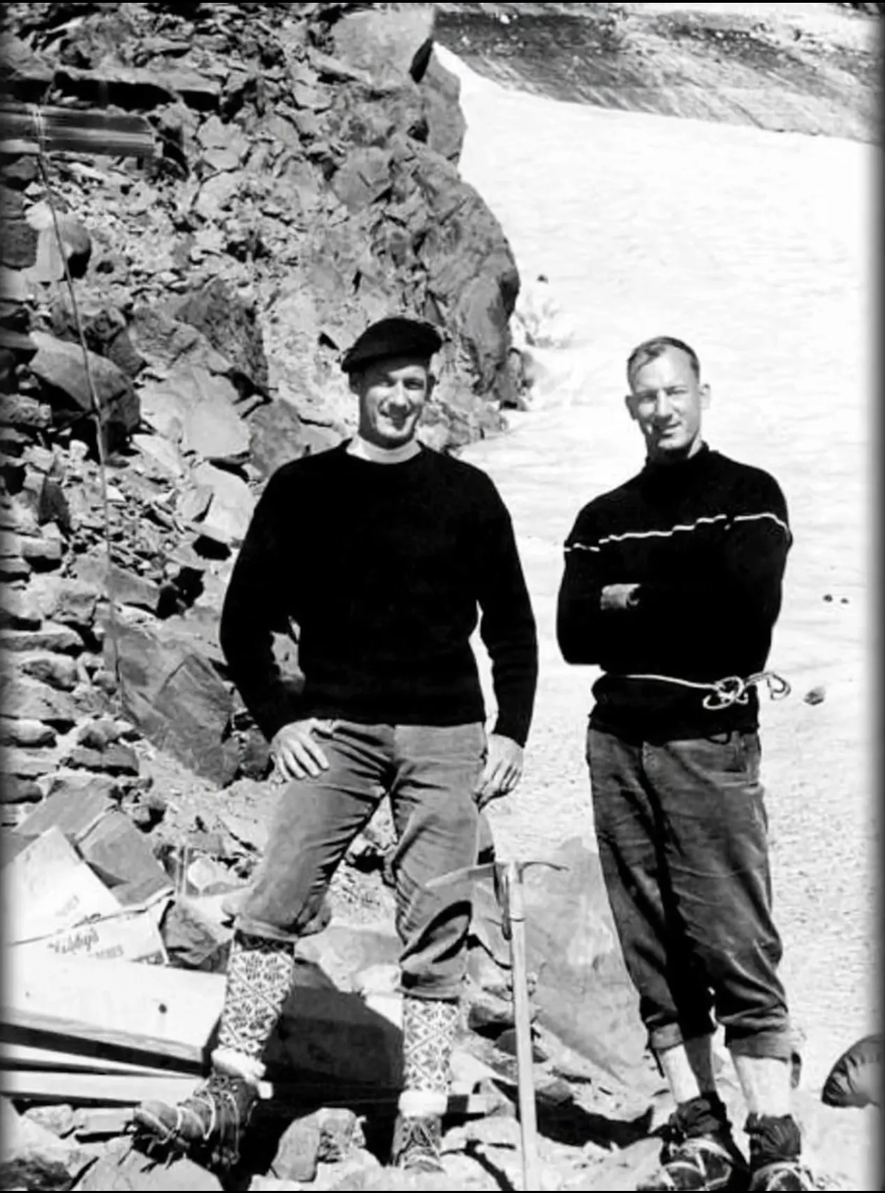 Black and white photo of two men in hiking boots and sweaters standing on rocky terrain with snow.
