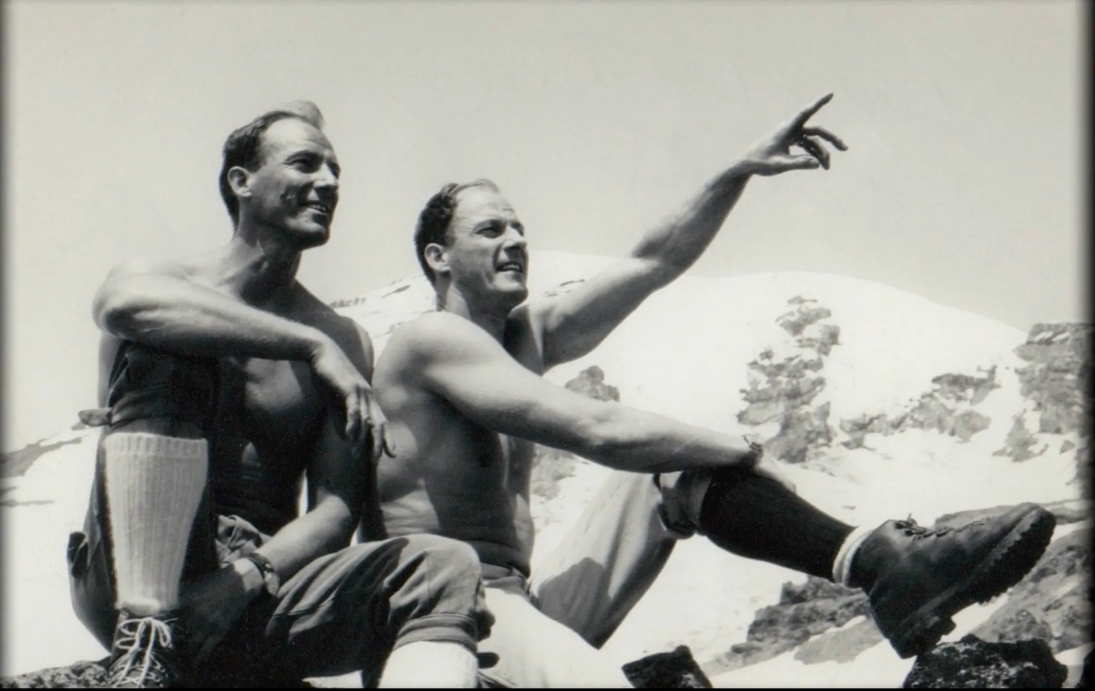 Two shirtless men sitting on rocks in a snowy mountain, one pointing into the distance.