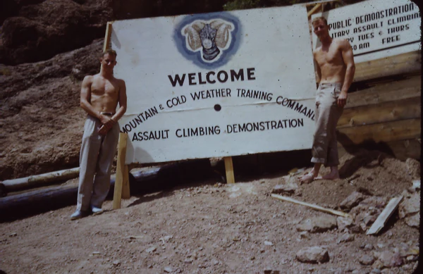Two shirtless men stand beside a sign welcoming to Mountain & Cold Weather Training Assault Climbing Demonstration.