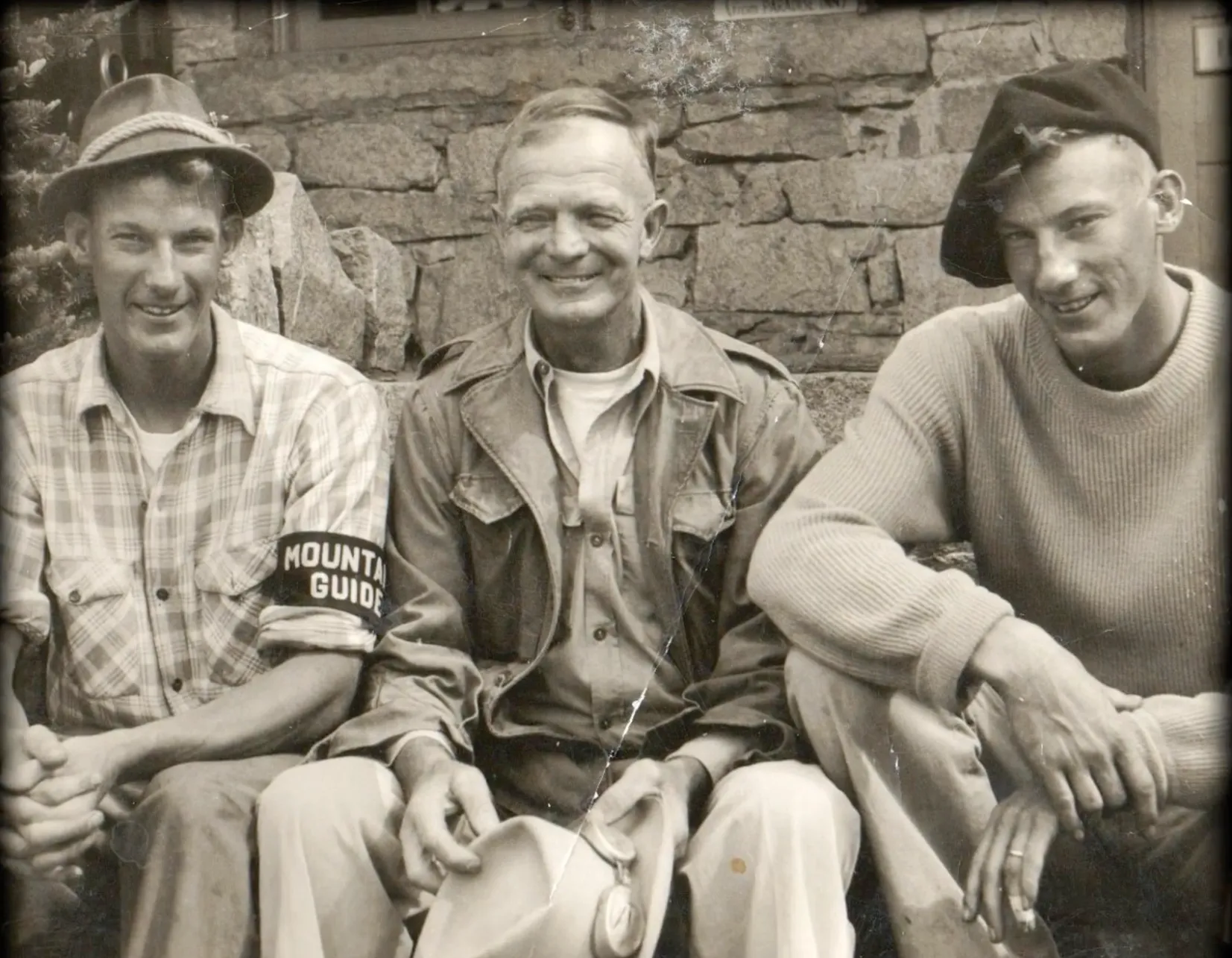 Three men sitting side by side in front of a stone wall, one wearing a Mountain Guide armband.