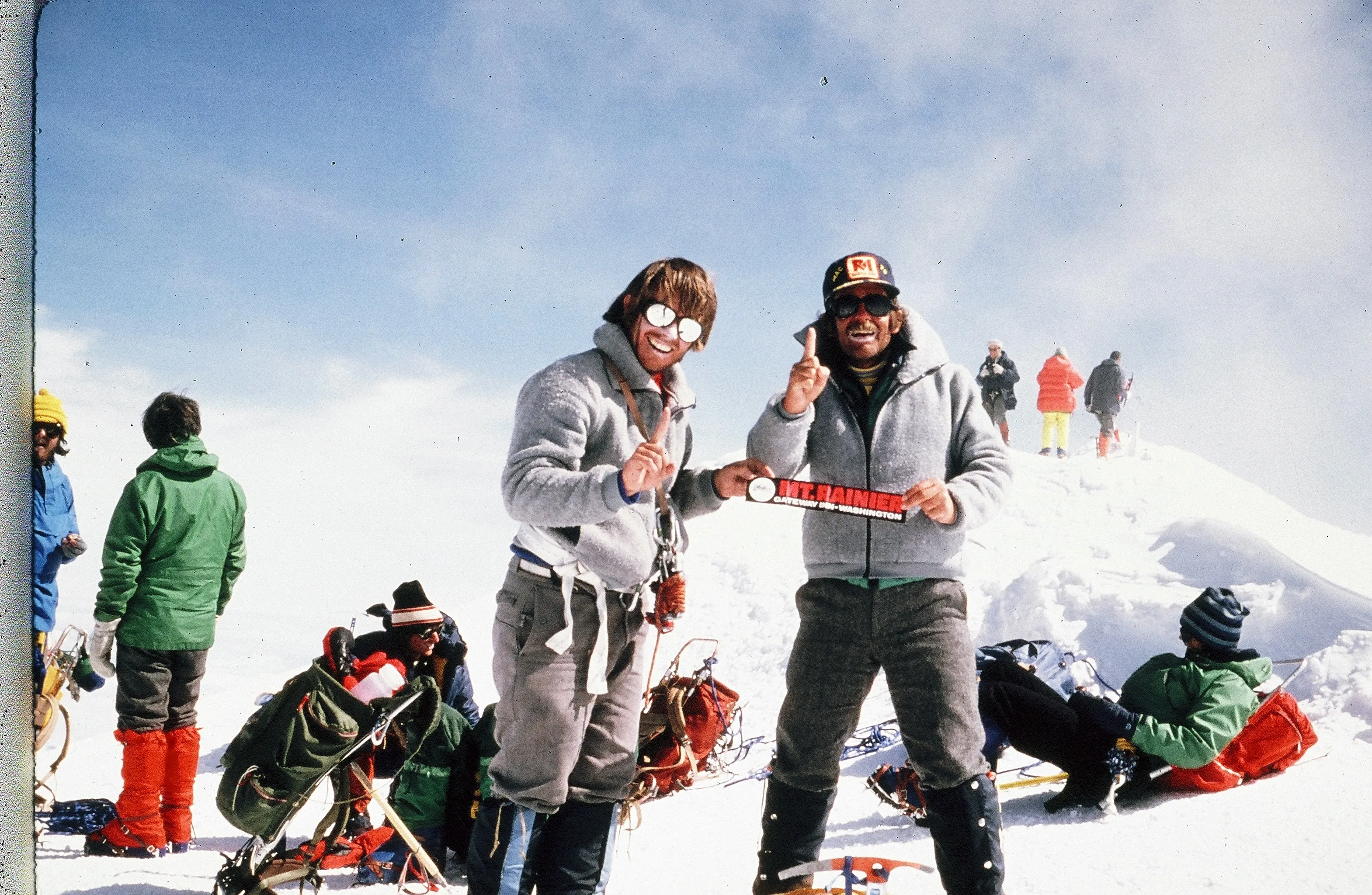 Two climbers in gray jackets holding a Mt. Rainier sticker, surrounded by others on snowy summit.