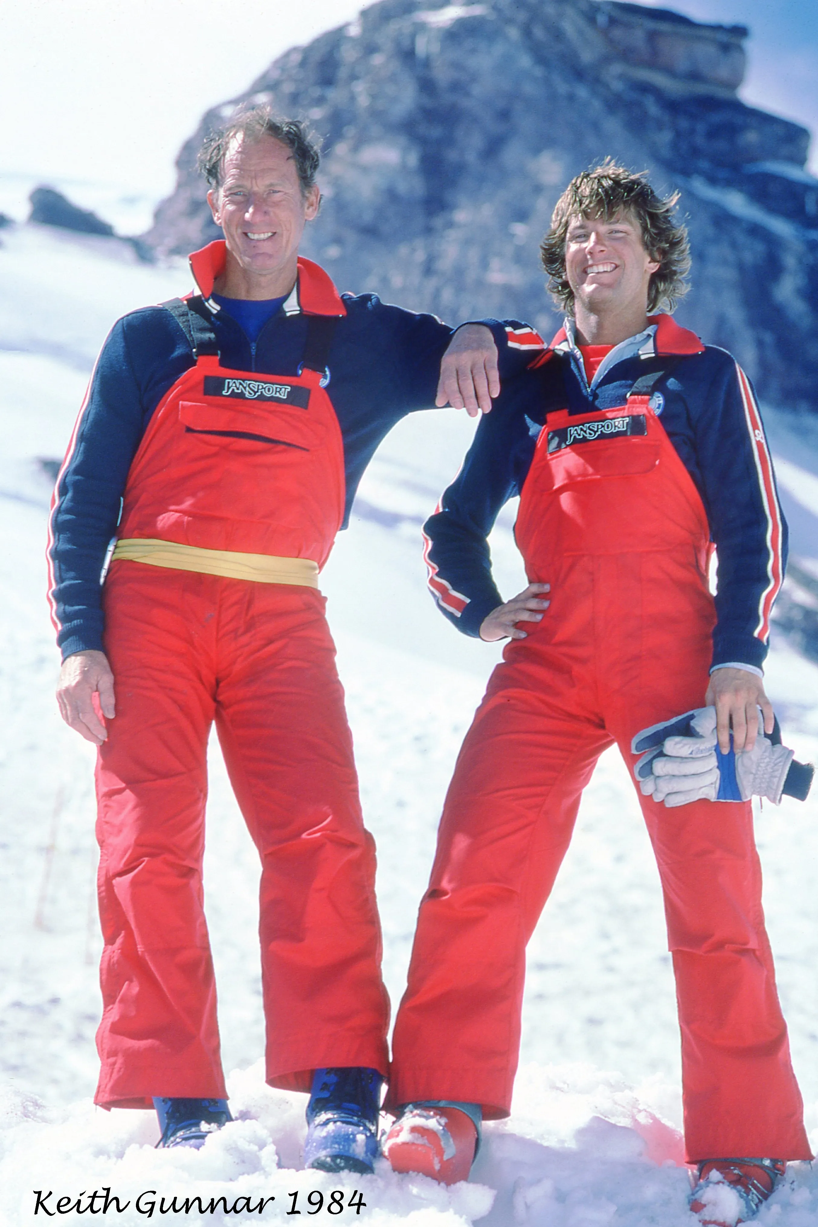 Two men in red Jansport ski overalls standing on snow with mountain rock behind them.