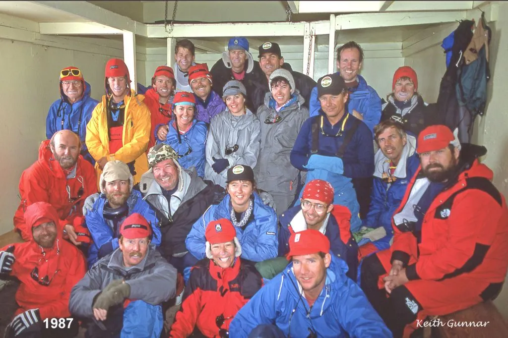 Group of 25 people dressed in colorful cold-weather gear posing inside a wooden shelter in 1987.