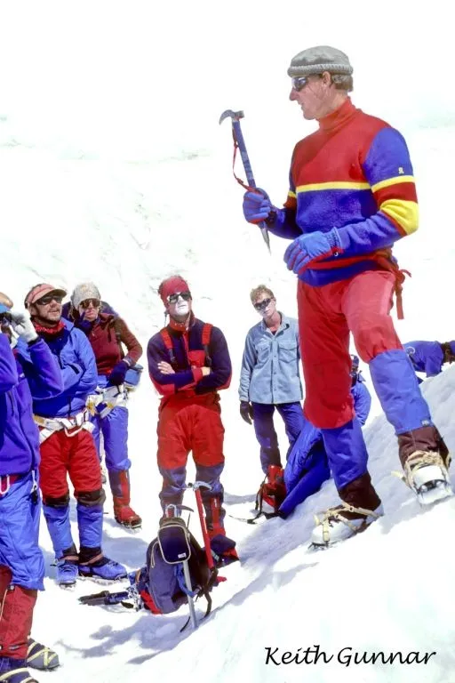 Man in colorful gear holding ice axe instructs group of climbers on snowy mountain slope.
