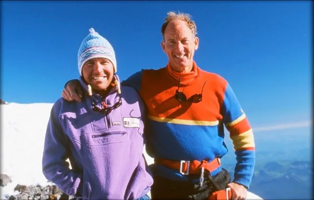 Two men smiling with arms around each other against a clear blue sky and snowy mountain background.