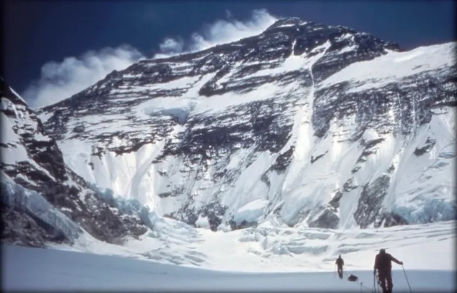 Two people skiing on snowy terrain towards a large snow-covered mountain with blue sky and clouds.