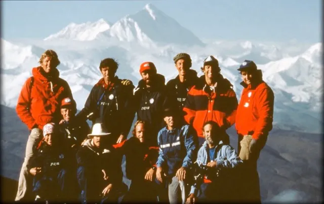 Group of climbers posing on a mountain with snowy peaks in the background under a clear sky.