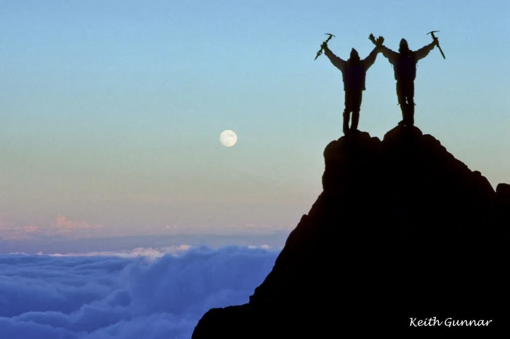 Silhouettes of two climbers holding ice axes standing atop a mountain with the moon nearby at dusk.