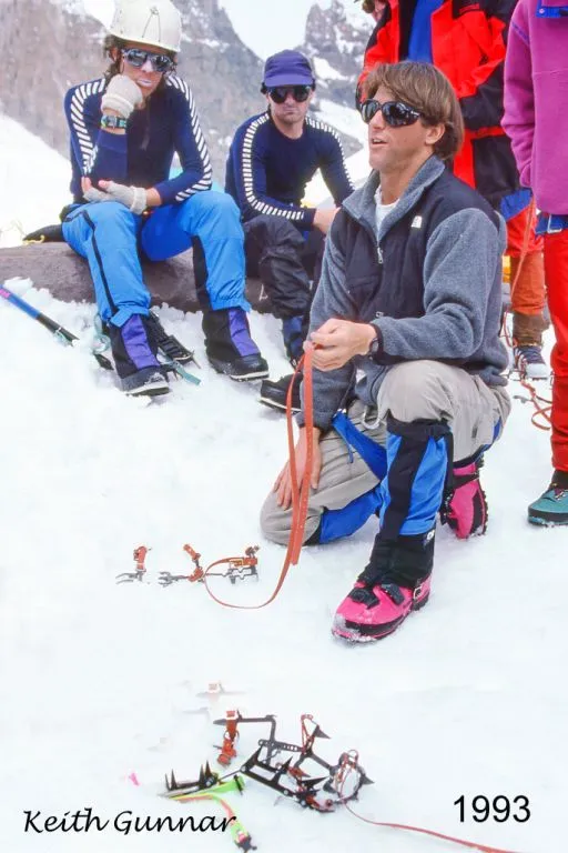 Man in sunglasses and winter gear kneels on snow teaching ice climbing to a group in mountainous terrain.