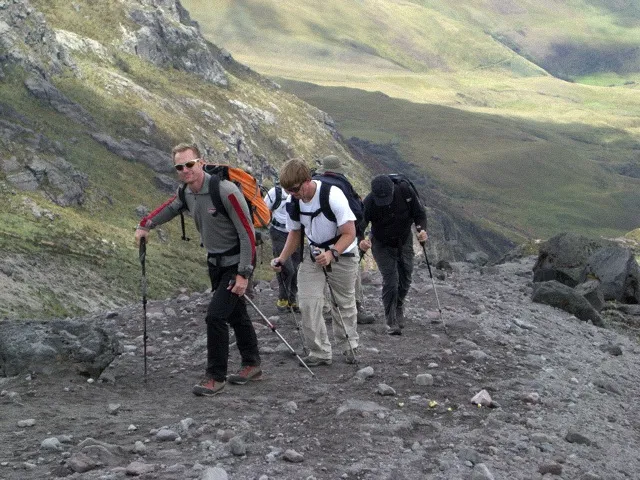 Four hikers with backpacks and walking sticks climbing a rocky mountain trail.