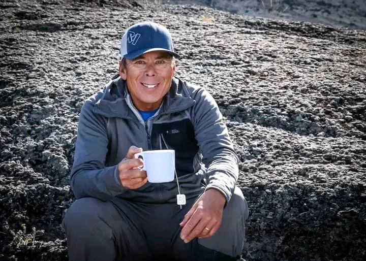 Man in gray jacket and cap sitting on rock, smiling, holding a white mug with a tea bag tag.