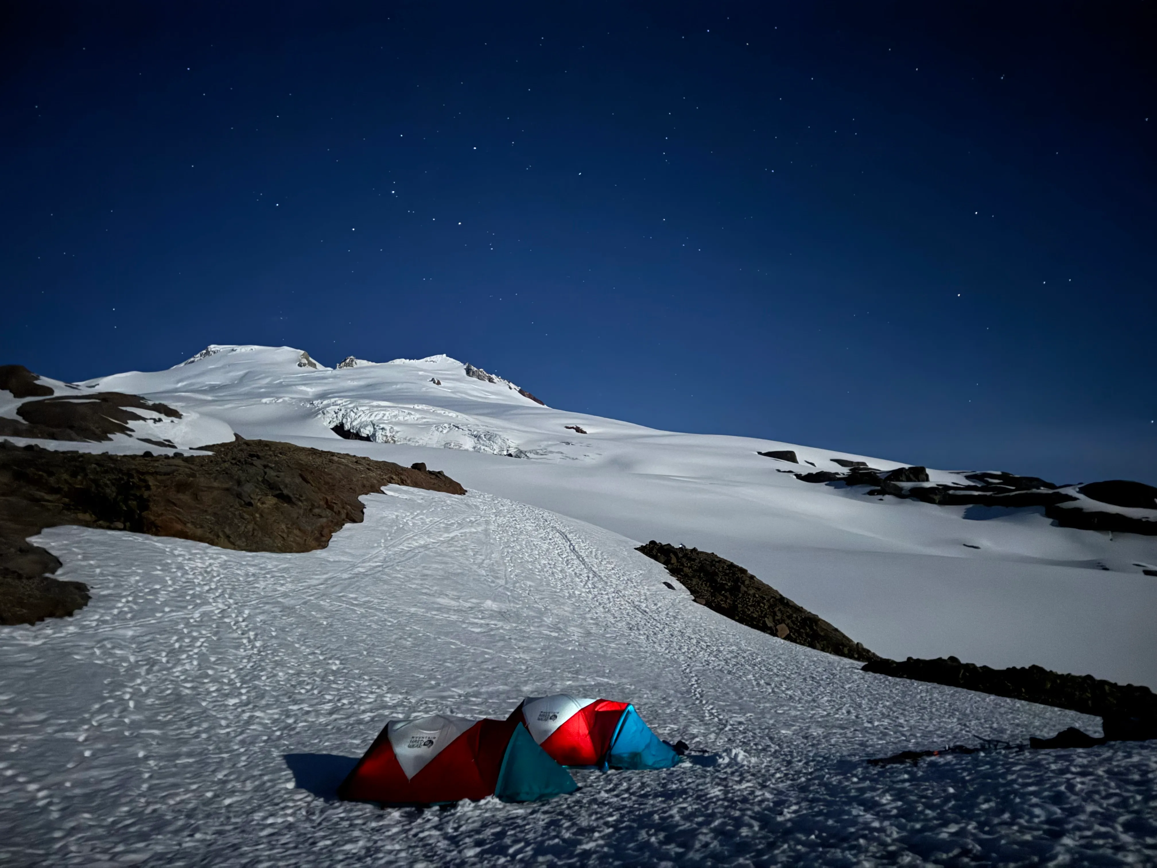 Three colorful tents pitched on snow at night under a starry sky with snowy mountain slopes.
