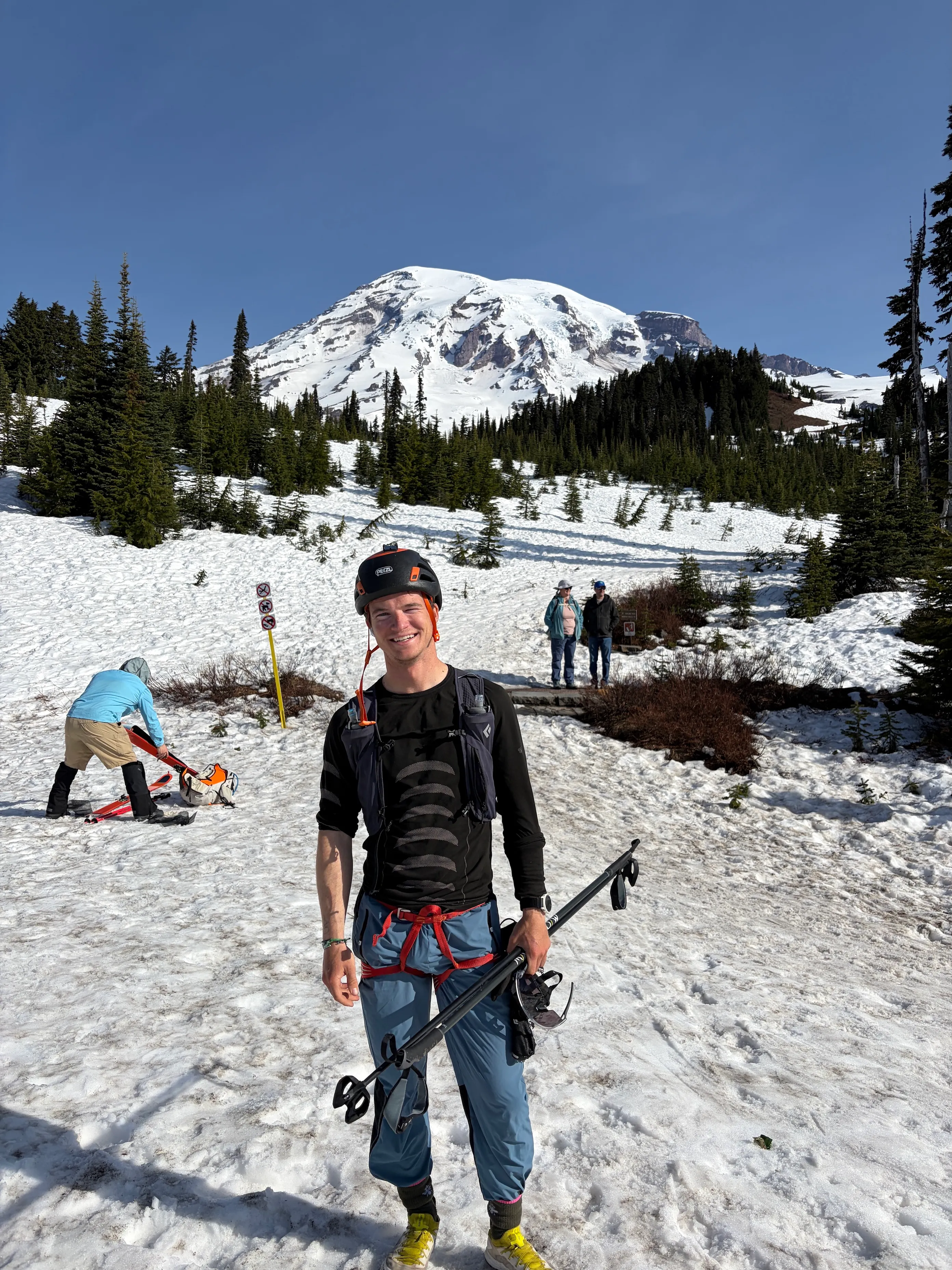 Smiling man in climbing gear holding ski poles with snowy mountain and trees in background.