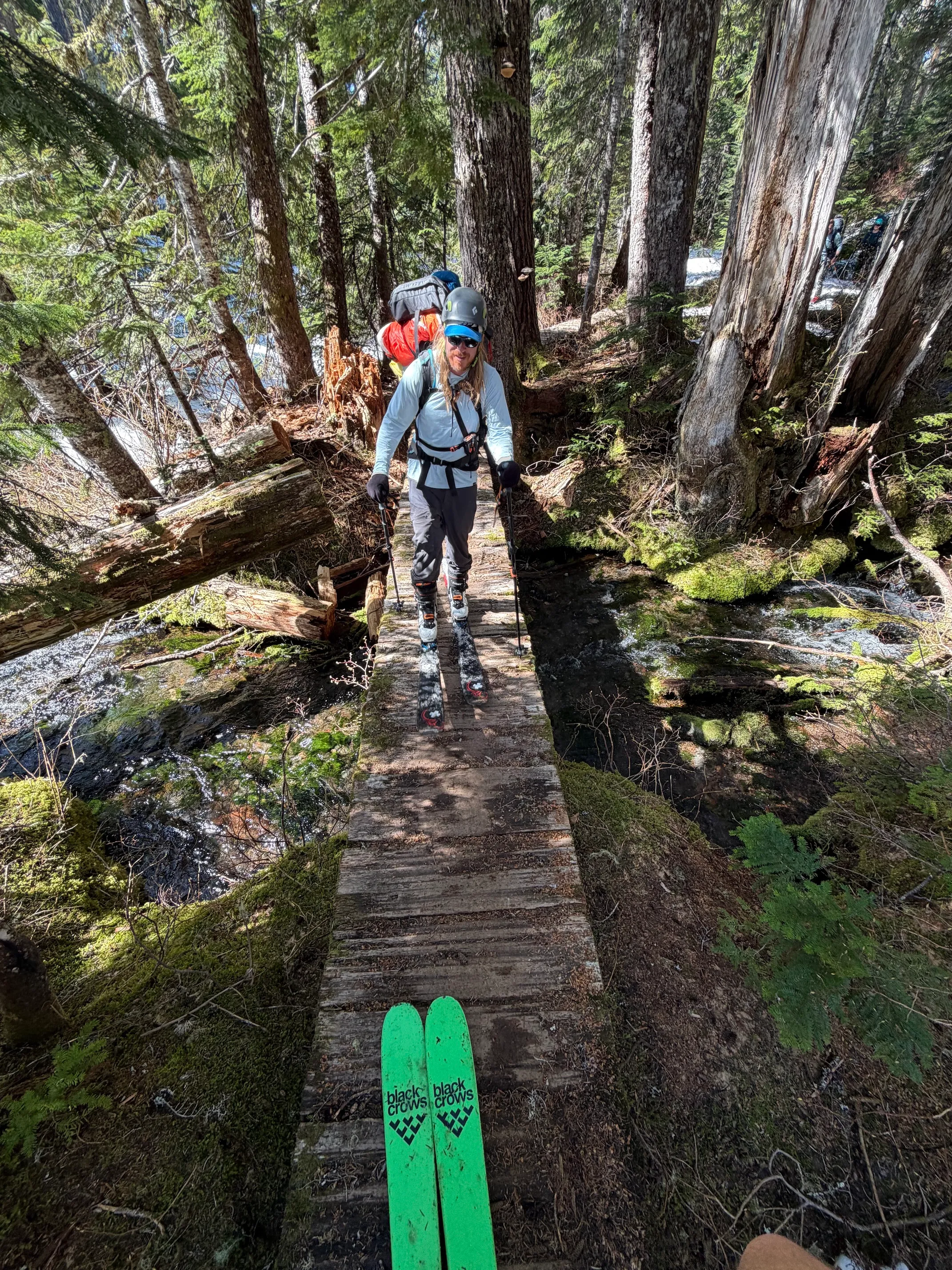 Skier with backpack crossing narrow wooden bridge in forest, visible green skis in foreground.