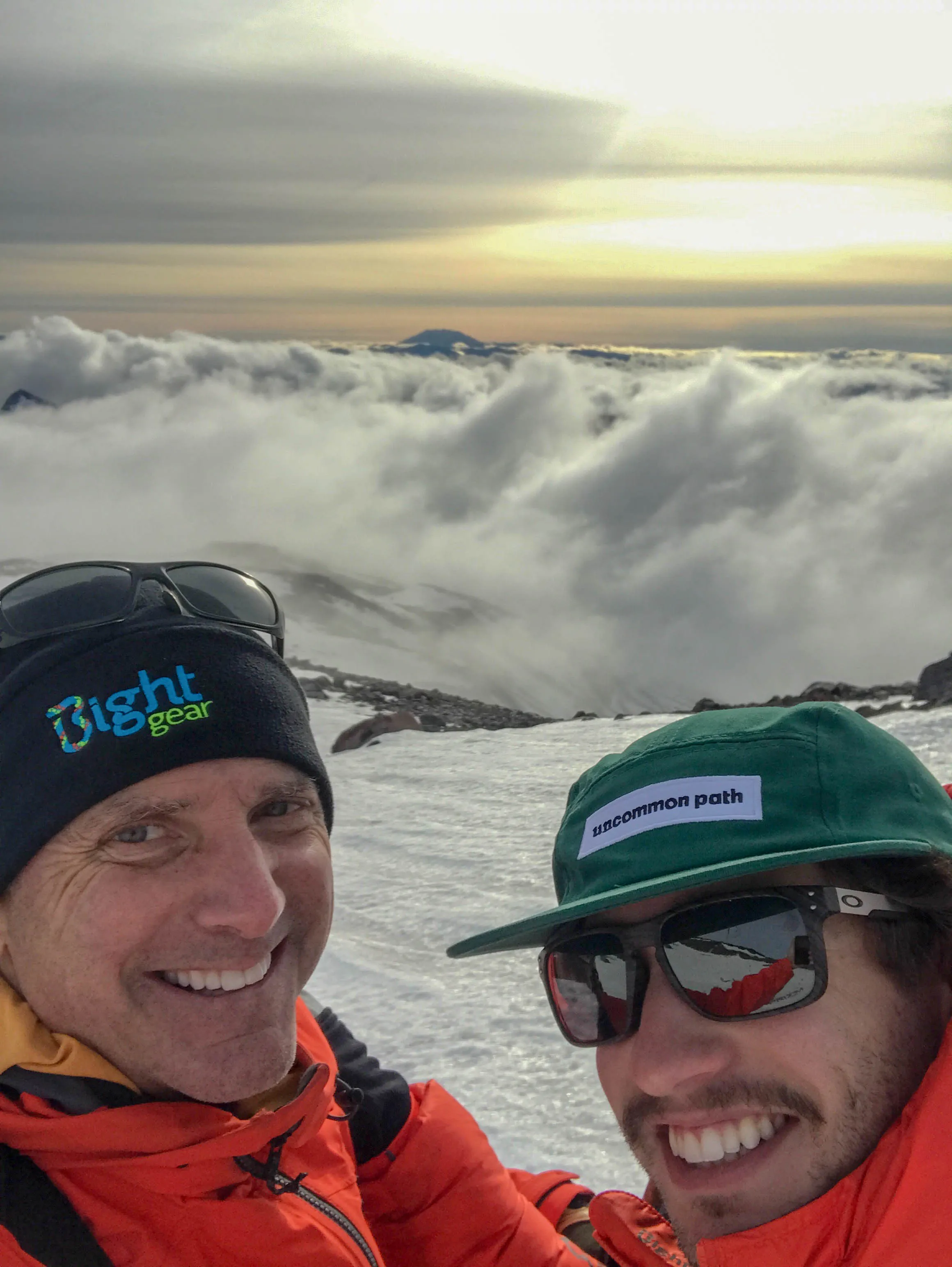 Two men in orange jackets and caps smiling on a snowy mountain with clouds and sunset behind them.