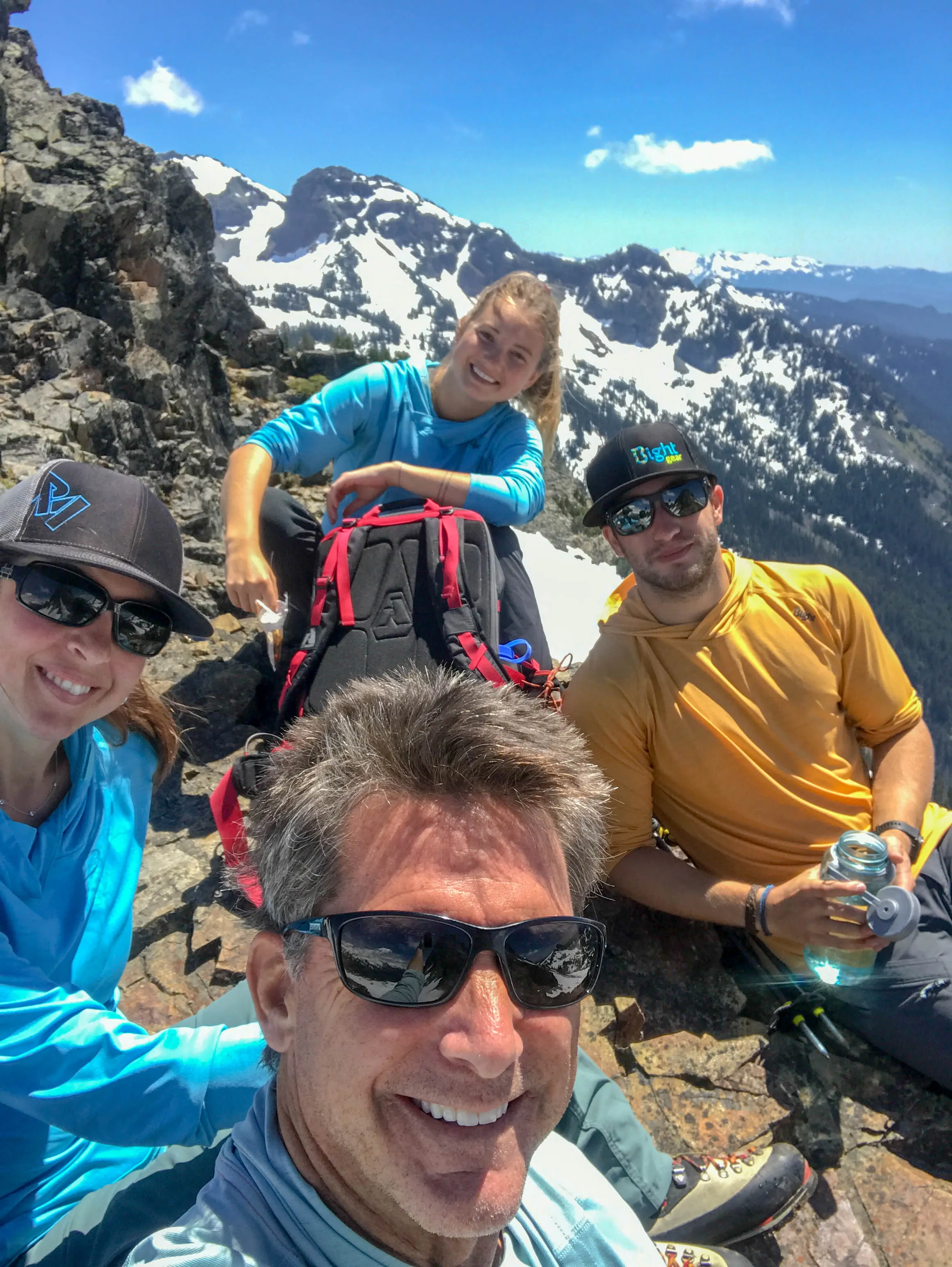 Four hikers resting on rocky summit with snow-capped mountains under blue sky behind them.