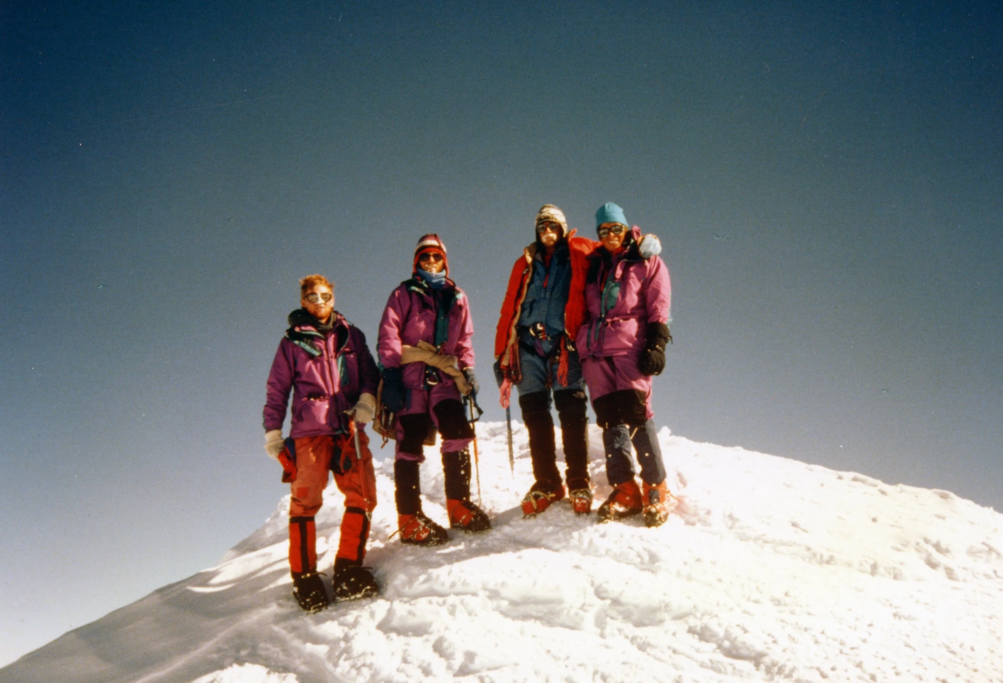 Four climbers in winter gear standing on a snow-covered mountain peak against a clear sky.